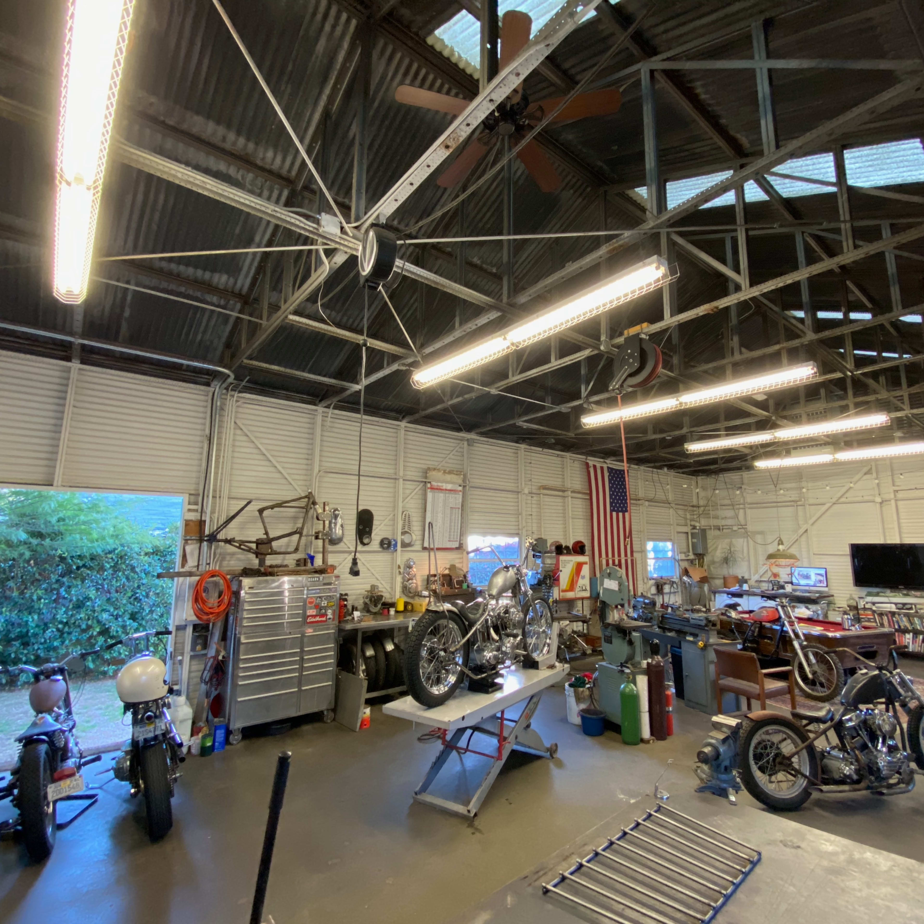 A workshop with multiple motorcycles and tools is illuminated by fluorescent lights under a high metal roof.