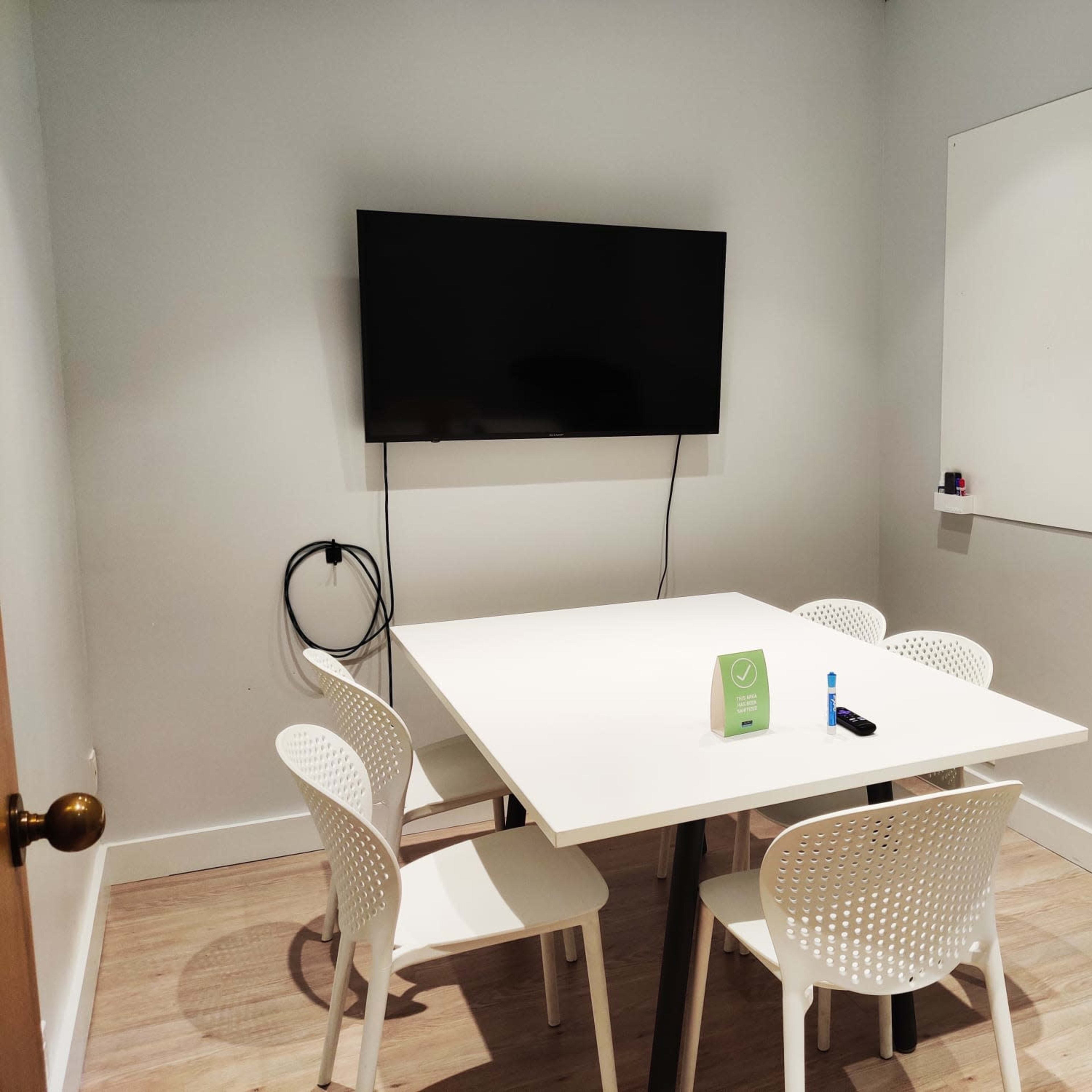 A small meeting room features a white table surrounded by four chairs, a television mounted on the wall, and a whiteboard.