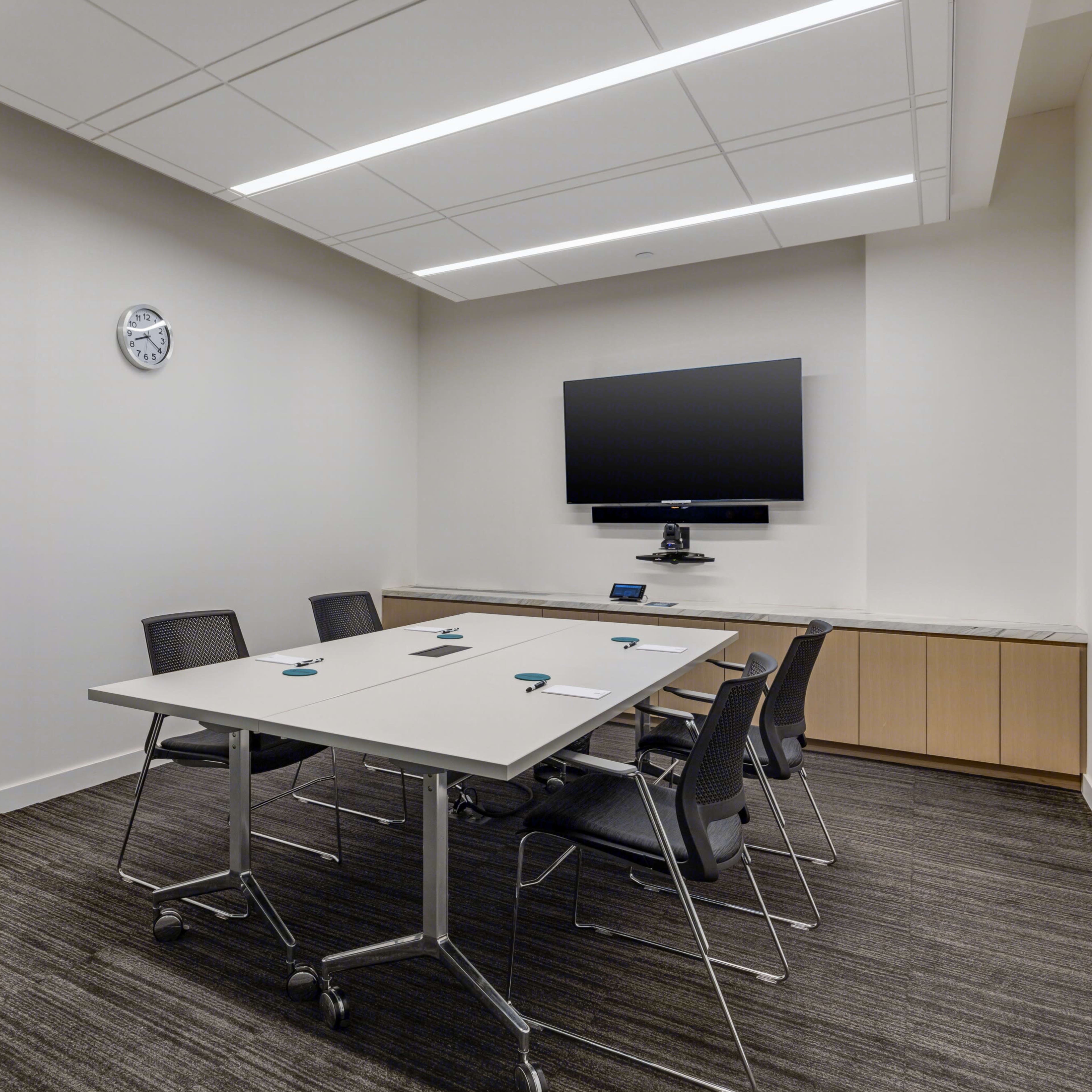 A small conference room features a rectangular table surrounded by chairs, with a wall-mounted screen and a clock visible.