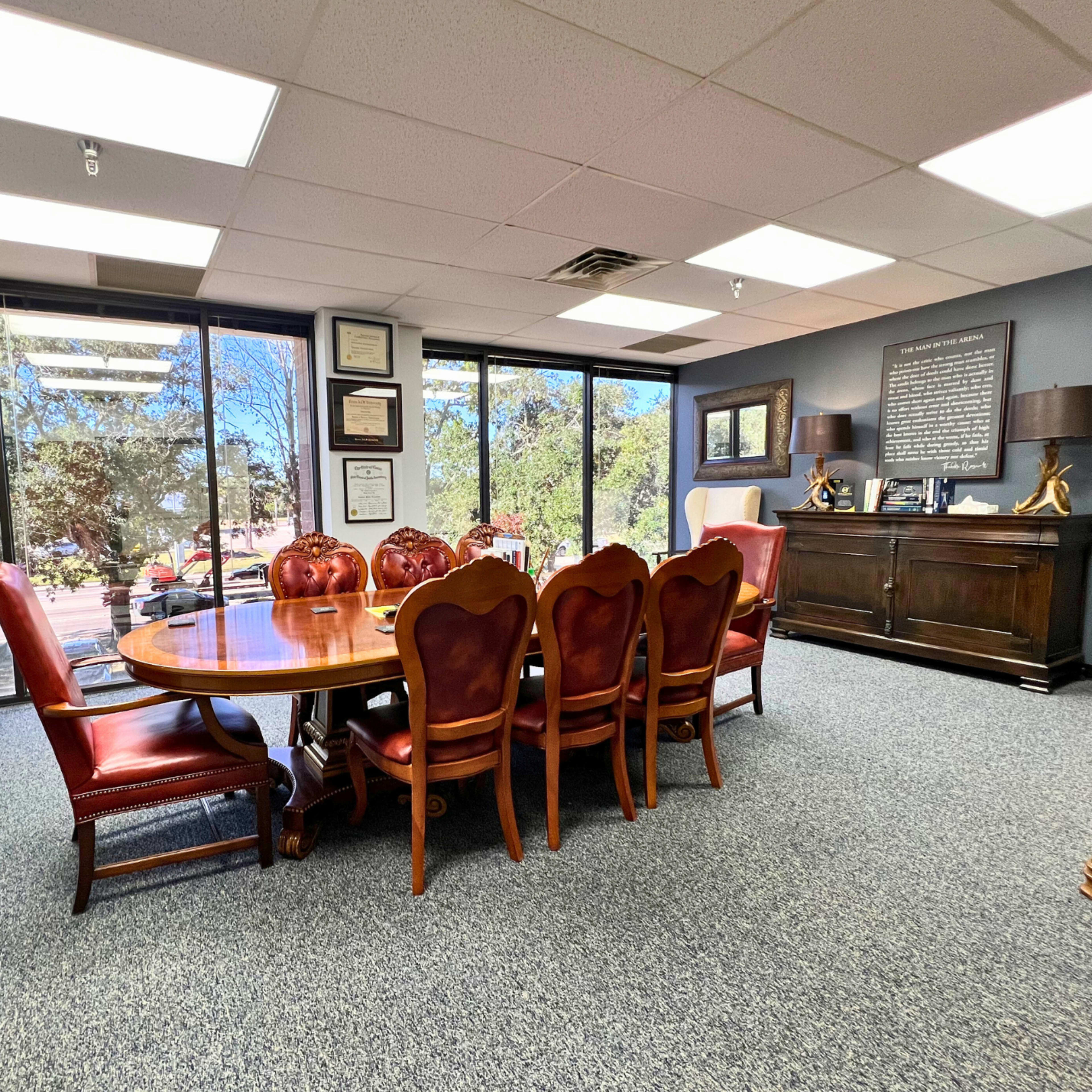 A conference room featuring a large wooden table surrounded by eight red upholstered chairs, with large windows providing natural light and a view of greenery outside.