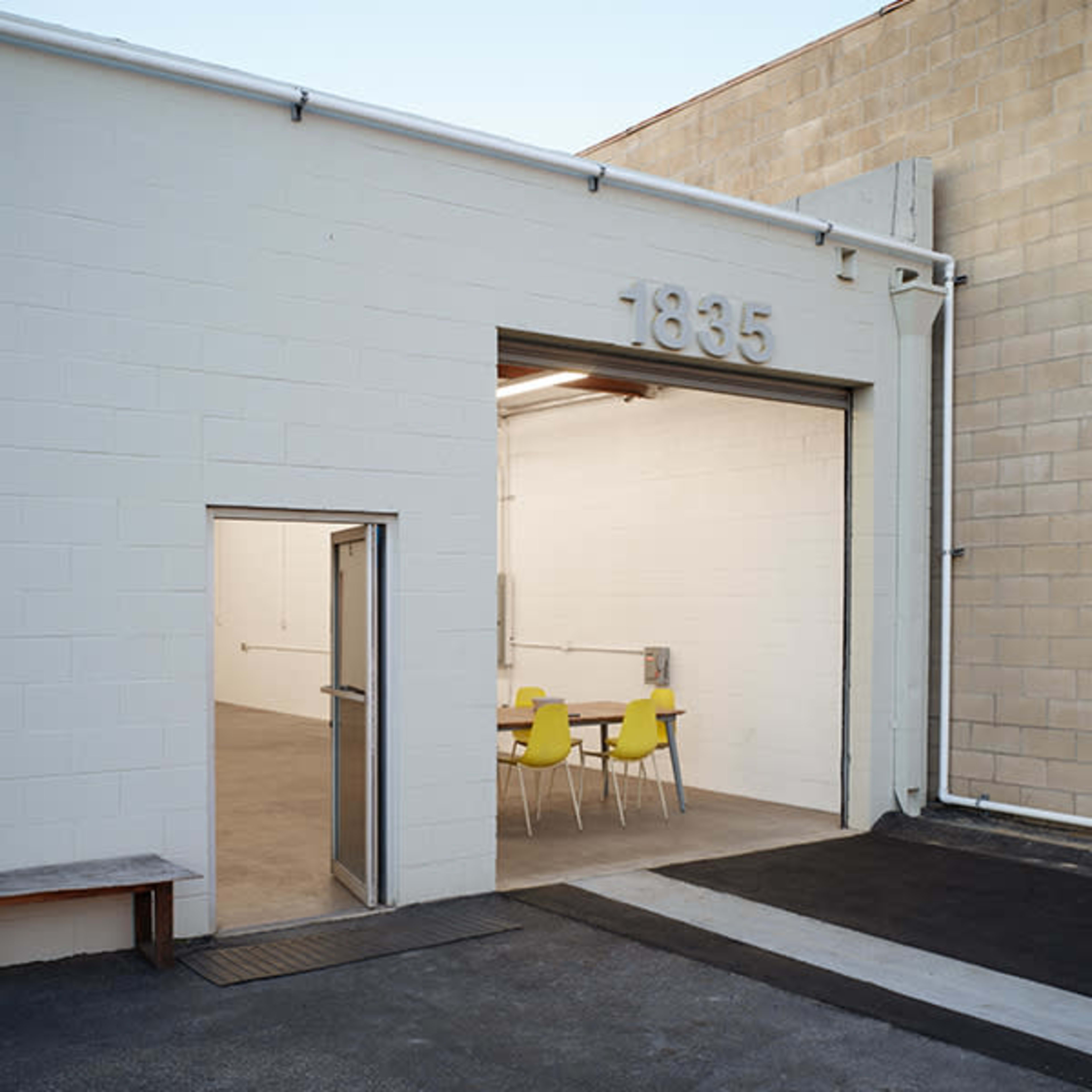 A simple, white warehouse exterior with the number 1835 displayed above an open entrance leading into a sparsely furnished interior with yellow chairs around a table.
