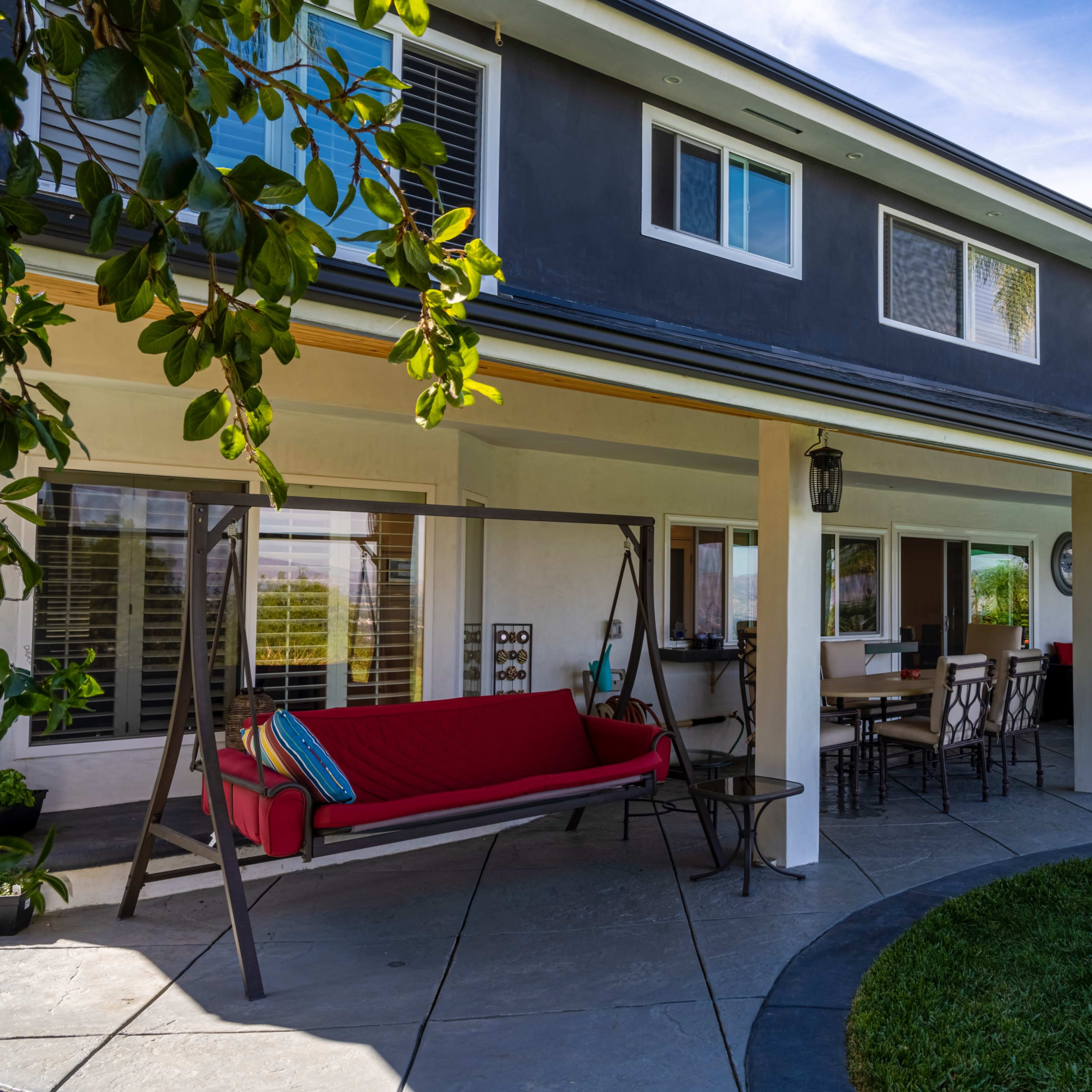 A modern two-story house with a covered patio, swing seat, and outdoor dining area surrounded by landscaped greenery.