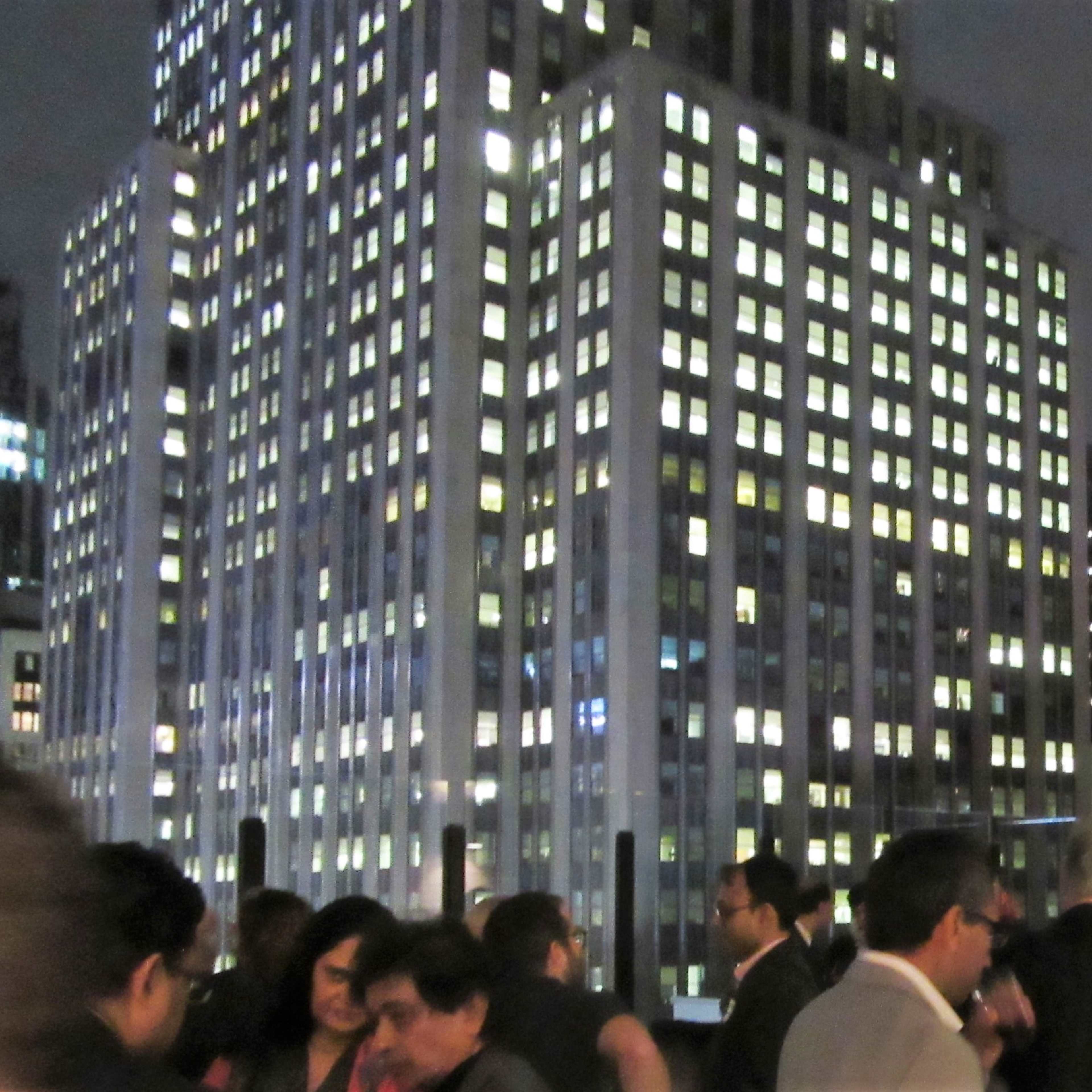 A bustling rooftop gathering at night, illuminated by city lights and surrounded by tall skyscrapers.