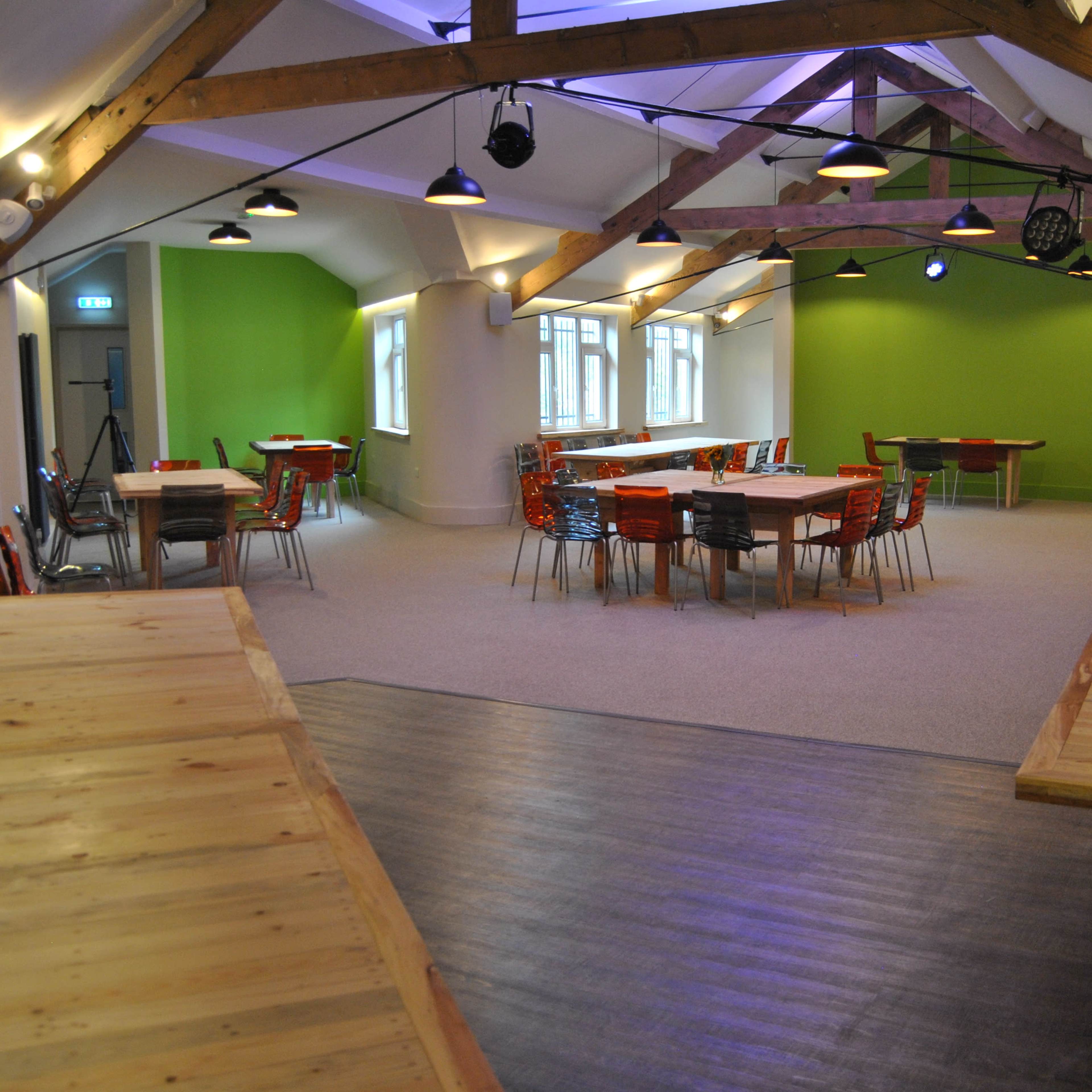 A spacious room with wooden tables and colorful chairs, featuring a green accent wall and exposed wooden beams.