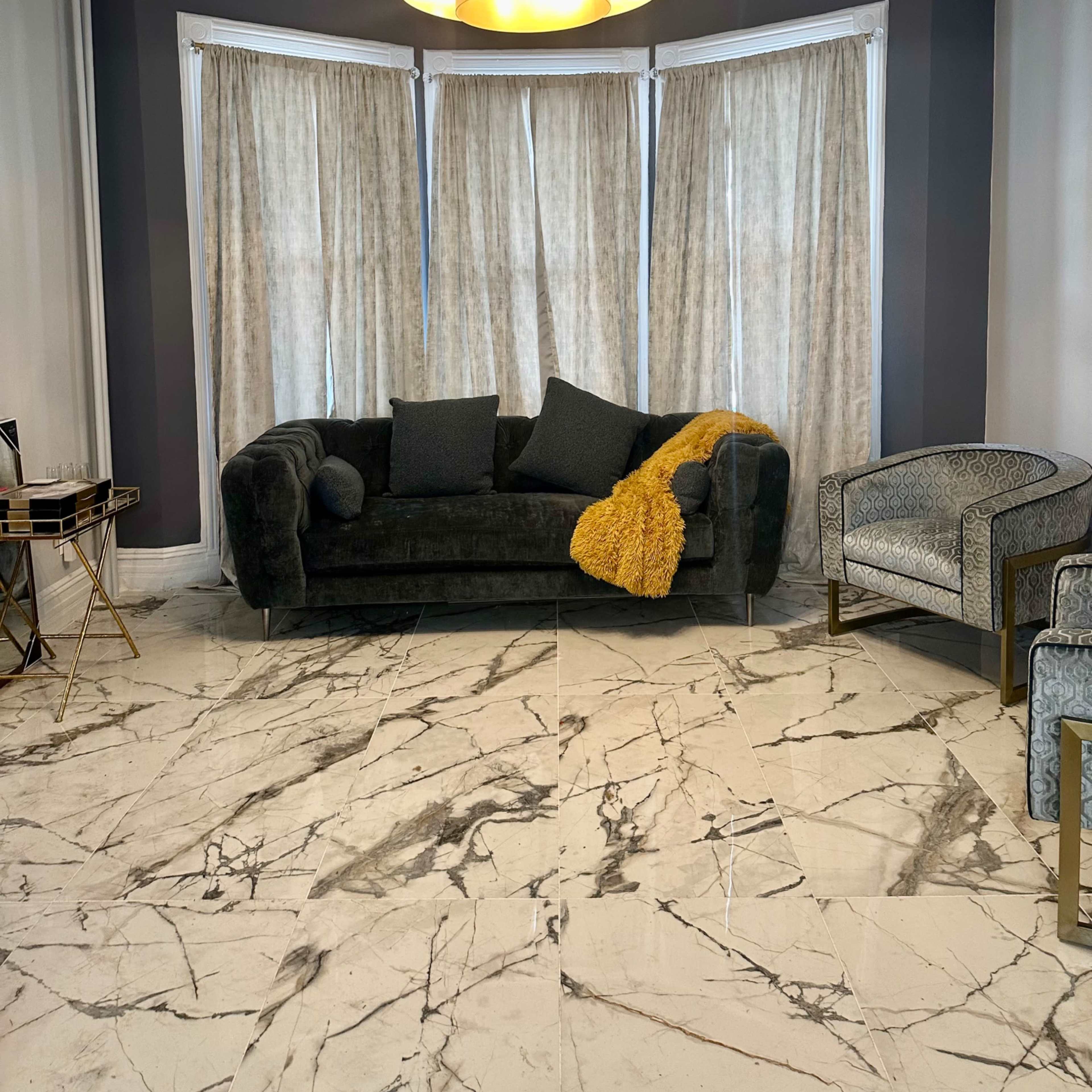 A dark couch and a patterned armchair are positioned in a room with marble flooring and large curtained windows.