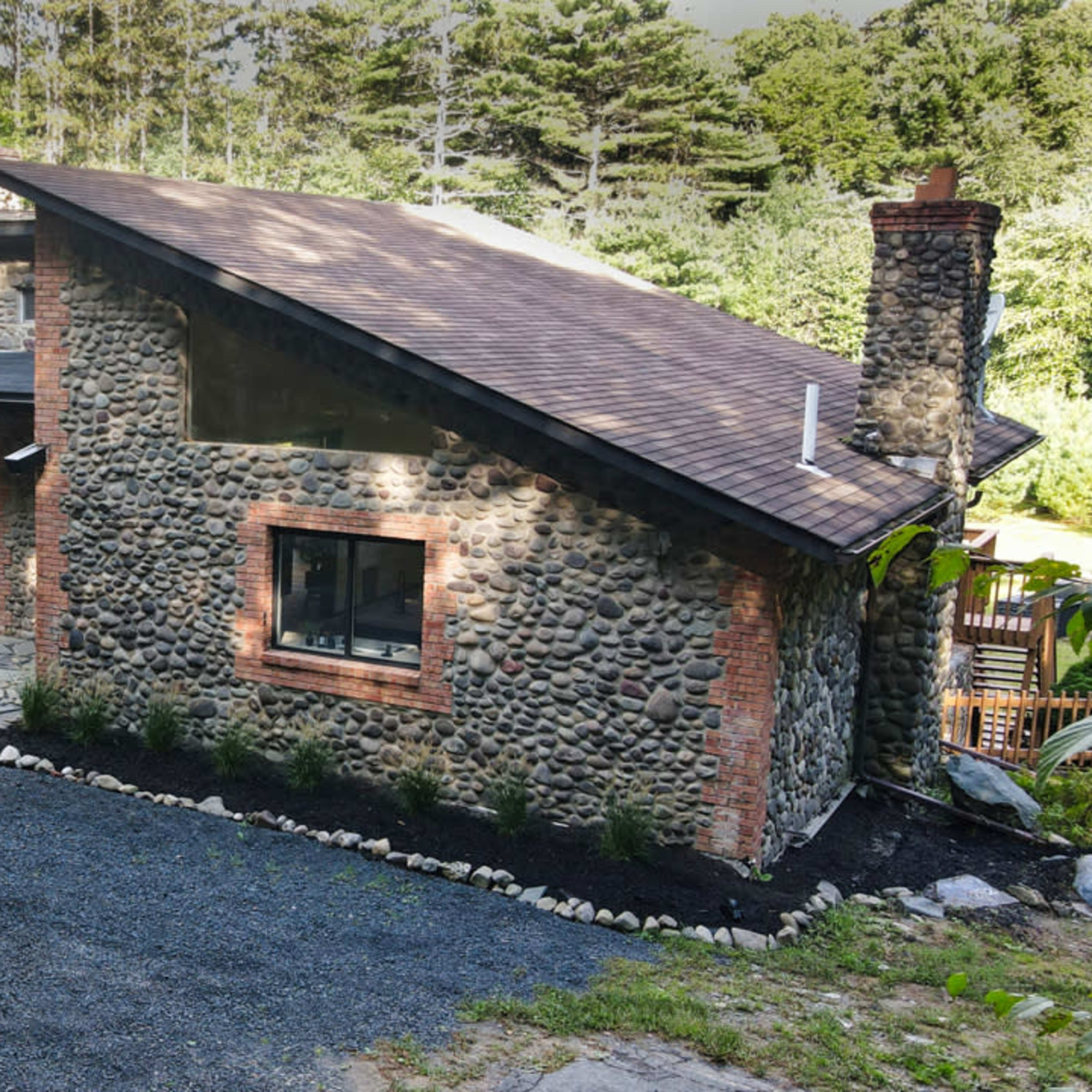 A stone and brick house with a sloped roof is set in a wooded area, featuring a landscaped pathway and outdoor seating.