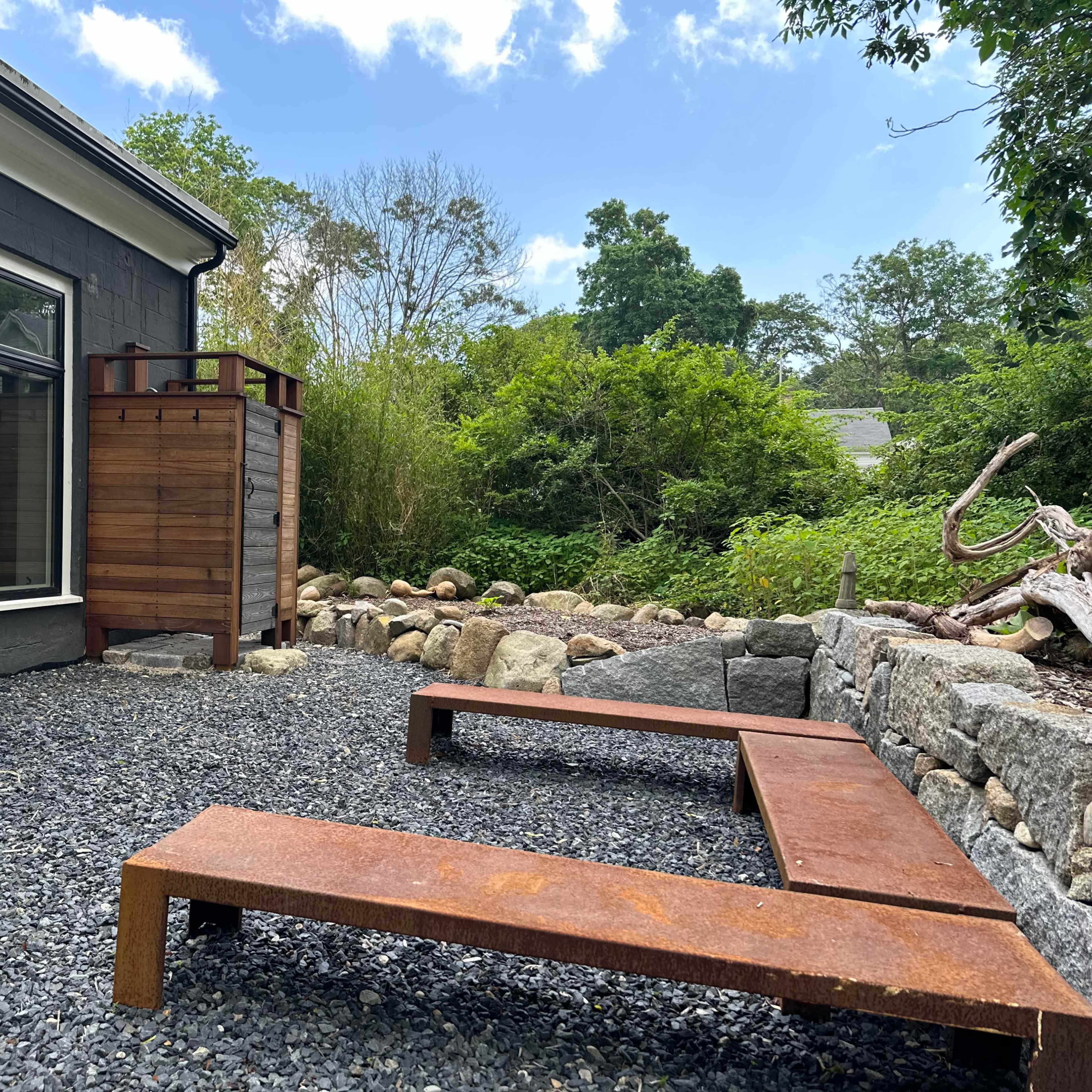 A landscaped outdoor area featuring a gravel surface, two rust-colored metal benches, and a wooden structure beside the gray house, surrounded by greenery and stone landscaping.