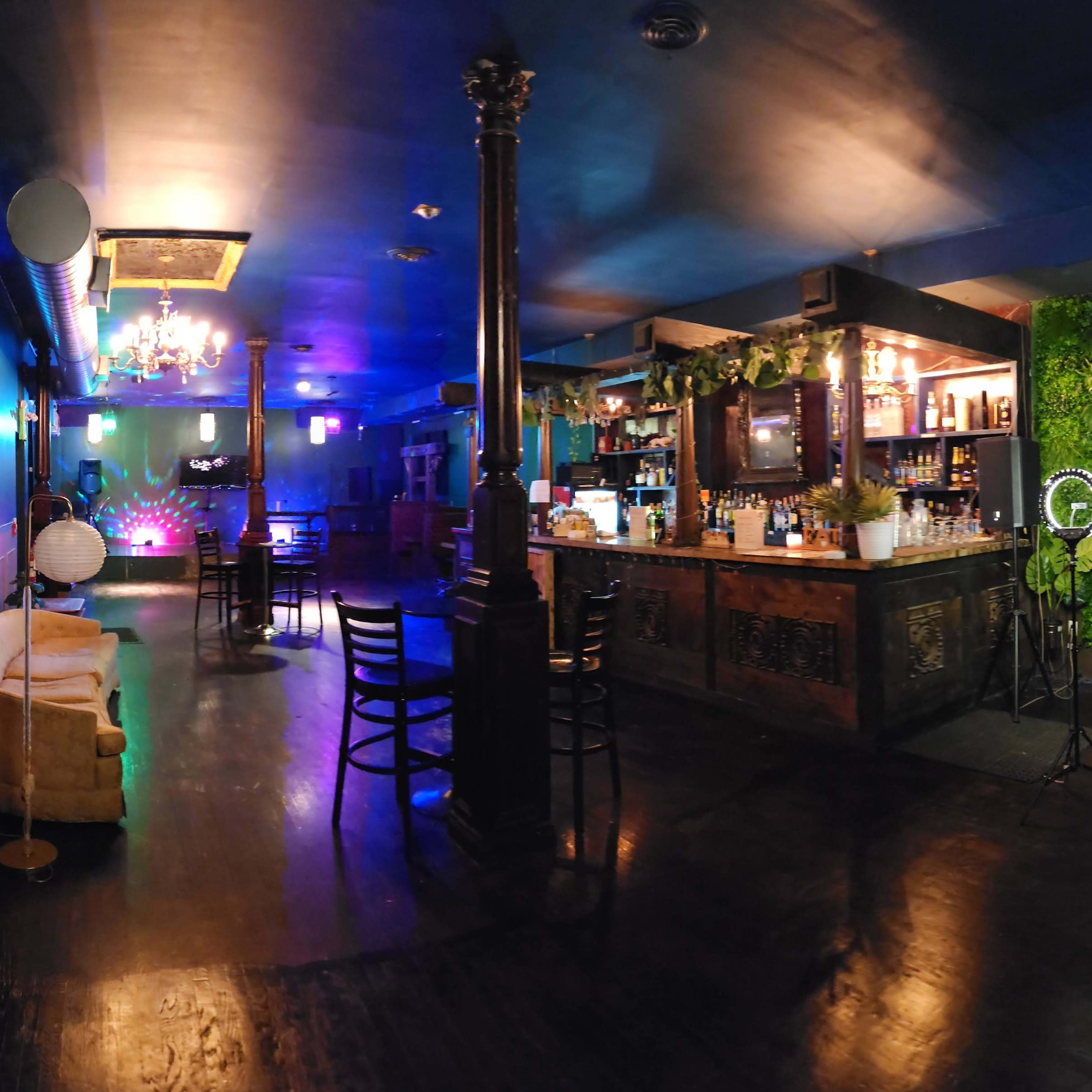 An indoor bar area with a wooden bar, lounge seating, a green wall, and dim lighting accented by colorful lights.