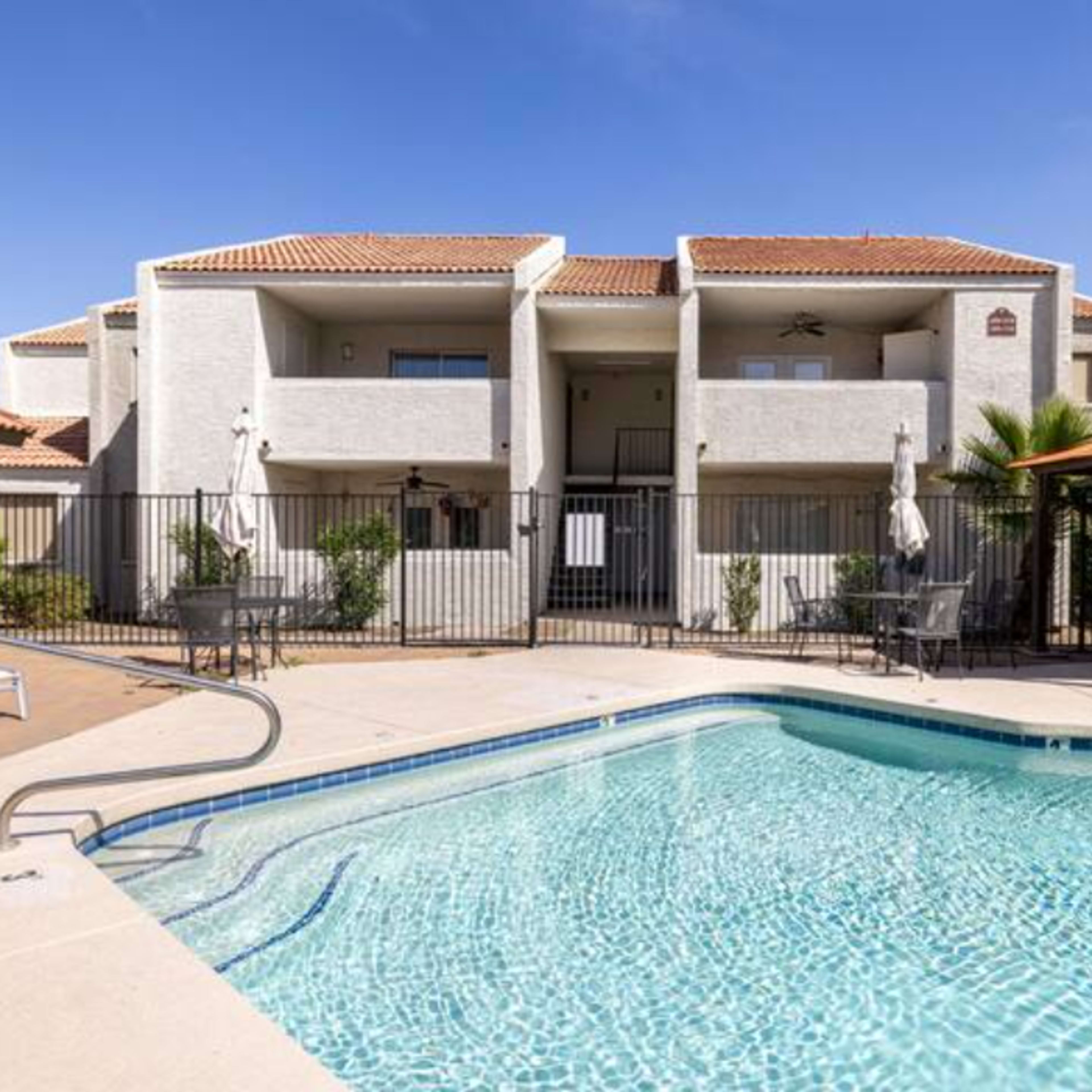 A clear swimming pool is surrounded by lounge chairs and palm trees, with an apartment building visible in the background.
