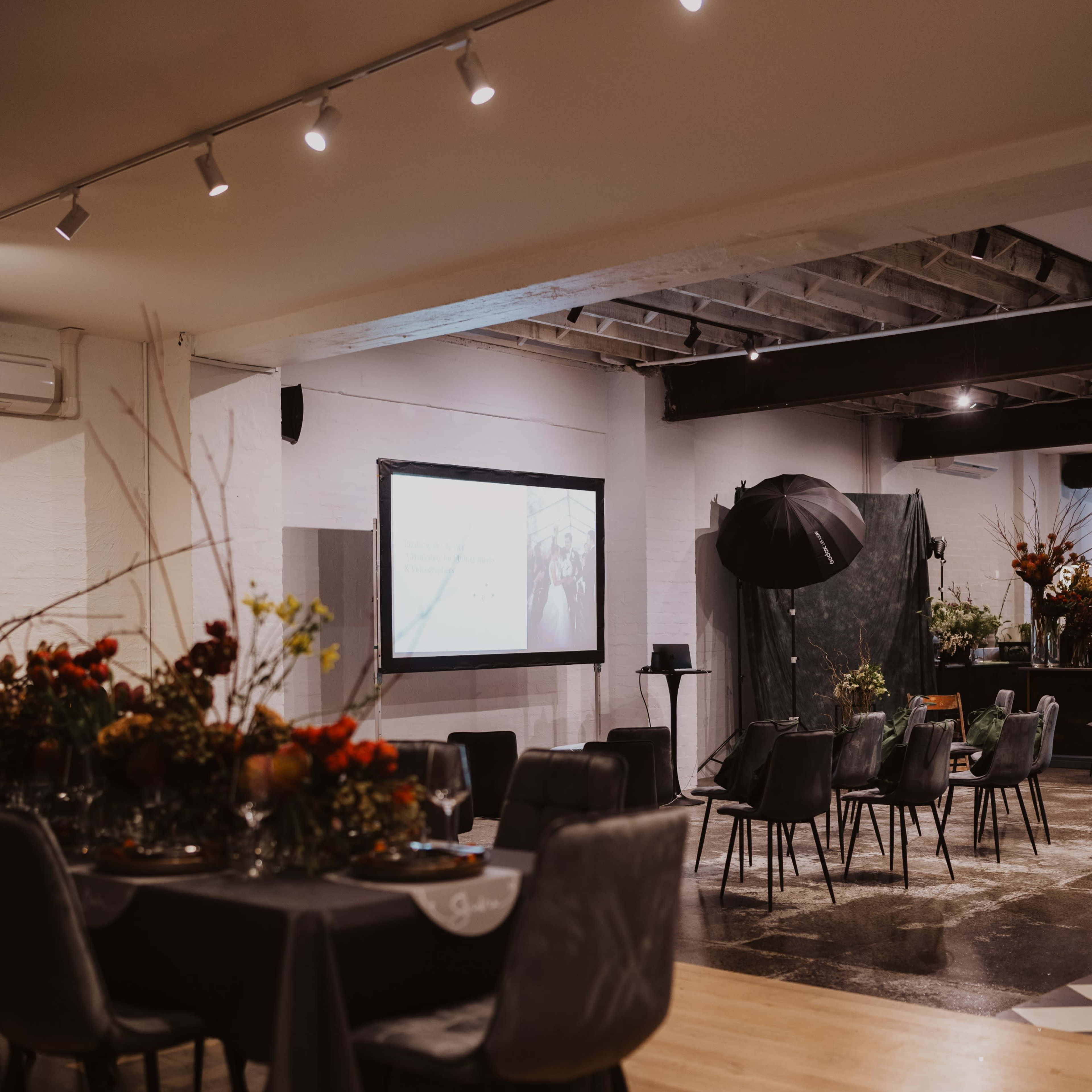 A spacious event venue features tables set for dining, a large screen for presentations, and decorative floral arrangements.