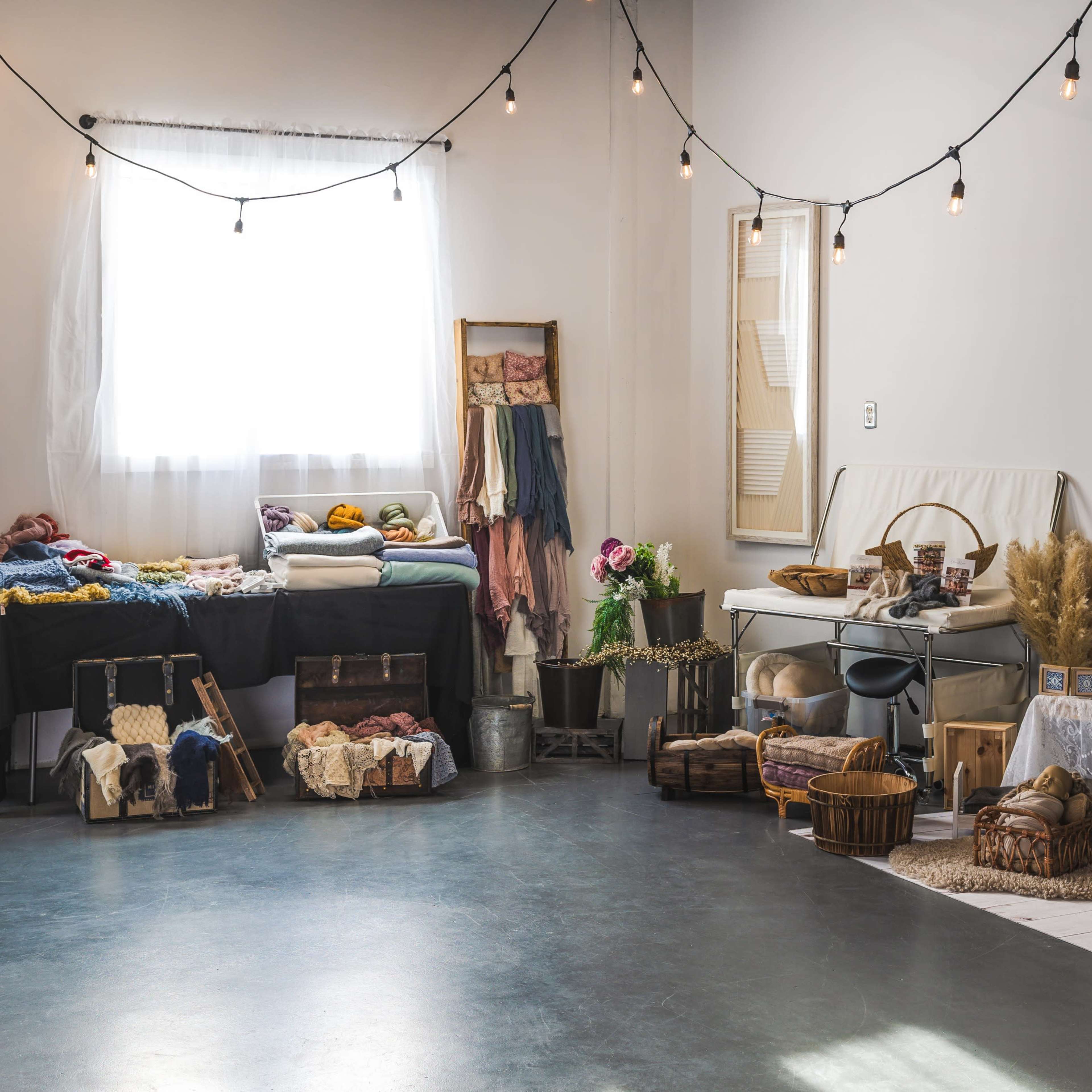 A brightly lit room features various textiles and baskets neatly arranged on tables and the floor, with string lights overhead and wooden walls in the background.