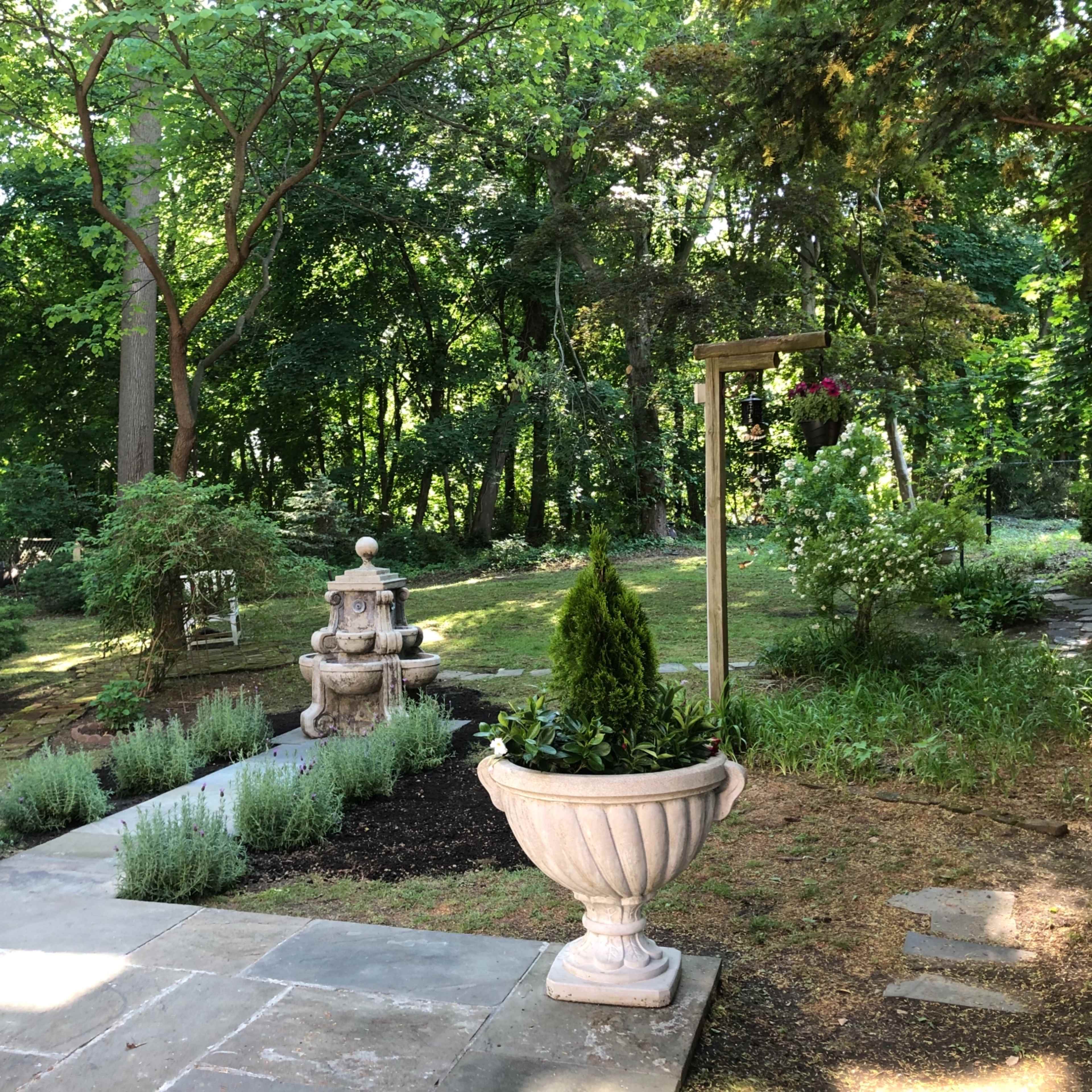 A stone pathway leads through a lush garden featuring a fountain and various plants surrounded by trees.