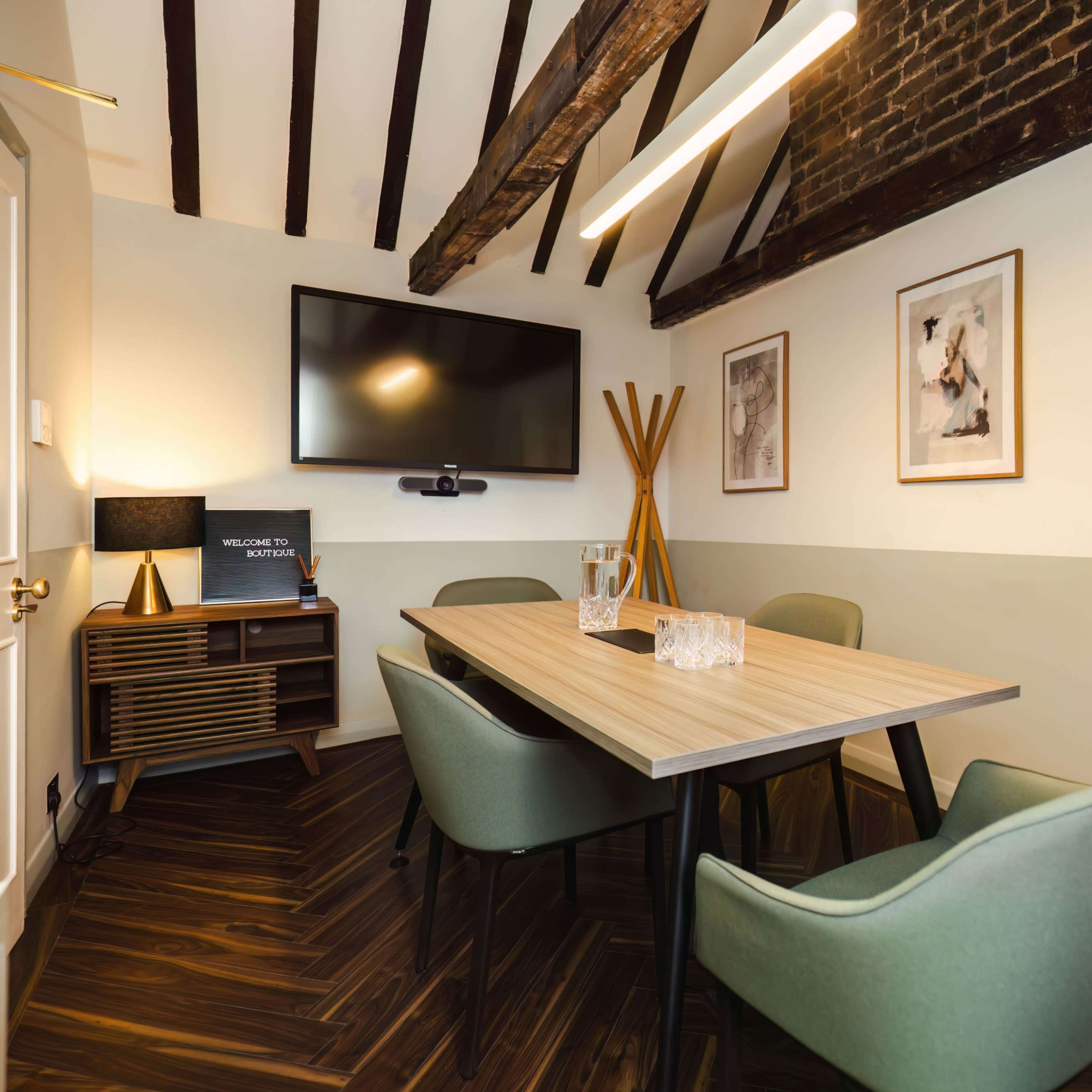 A conference room features a wooden table surrounded by green chairs, with a television mounted on the wall and framed artwork displayed.