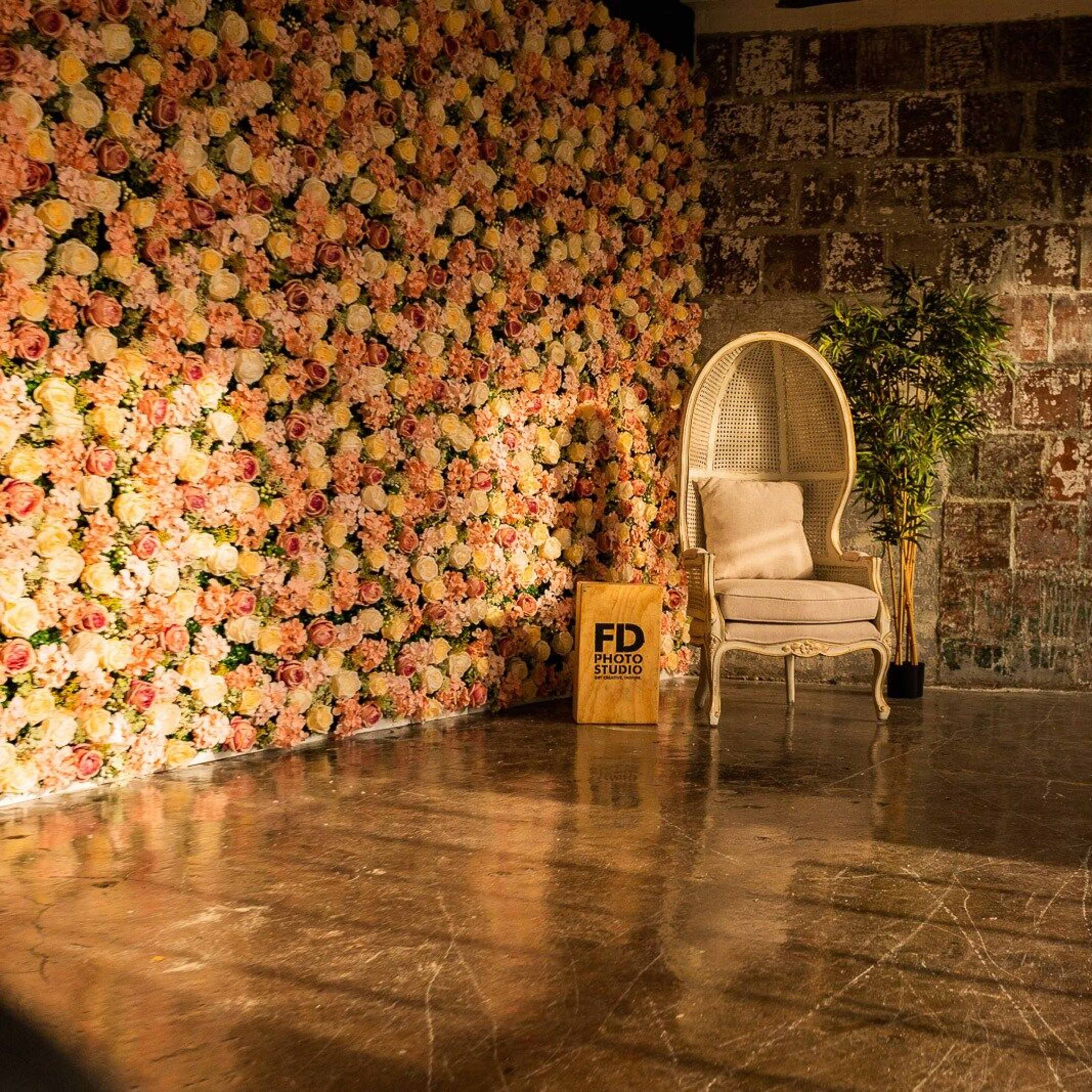 A vintage white chair positioned in front of a wall adorned with a colorful flower arrangement, set in an industrial space with a concrete floor.
