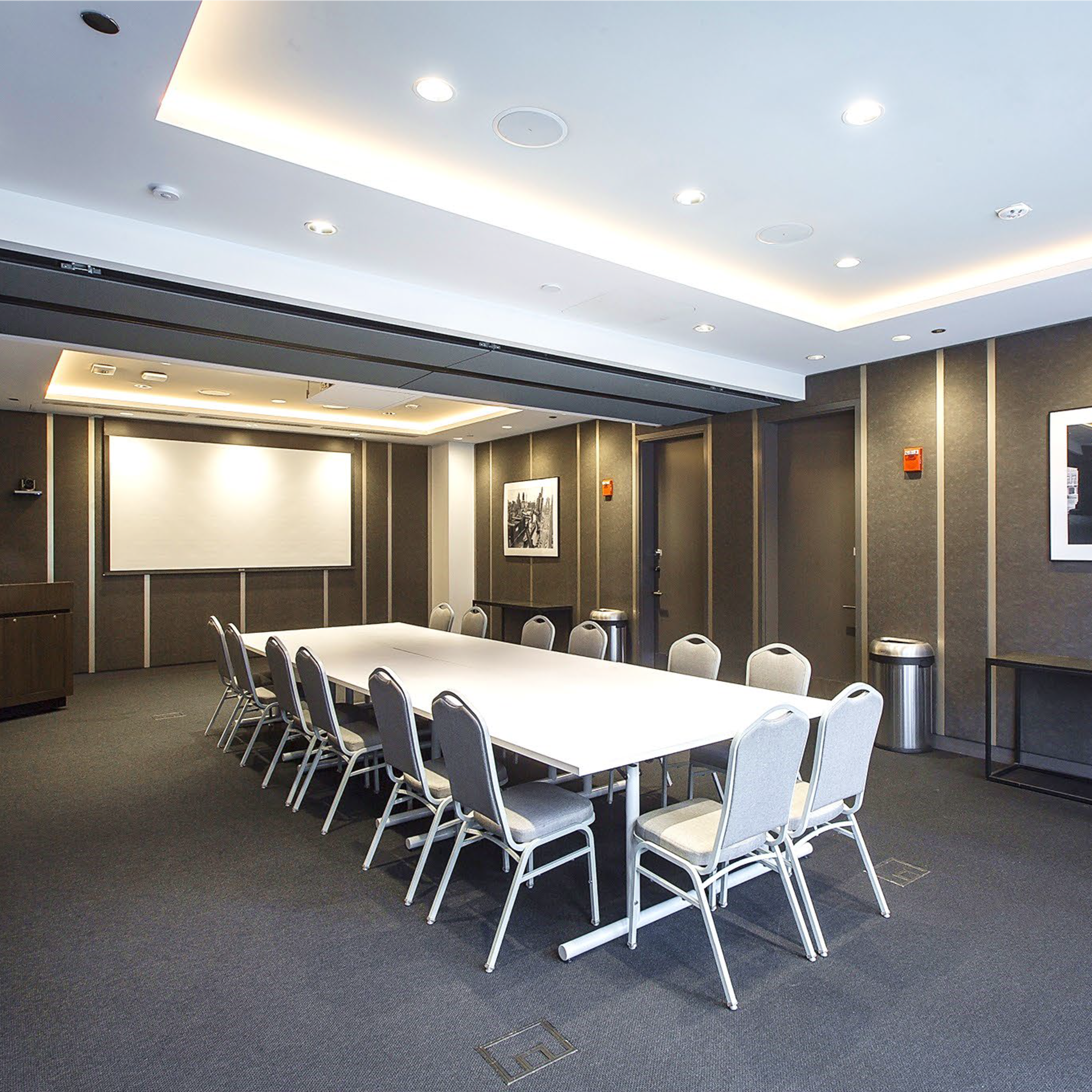 A modern conference room featuring a long white table surrounded by silver chairs, with a projector screen and framed photographs on the walls.