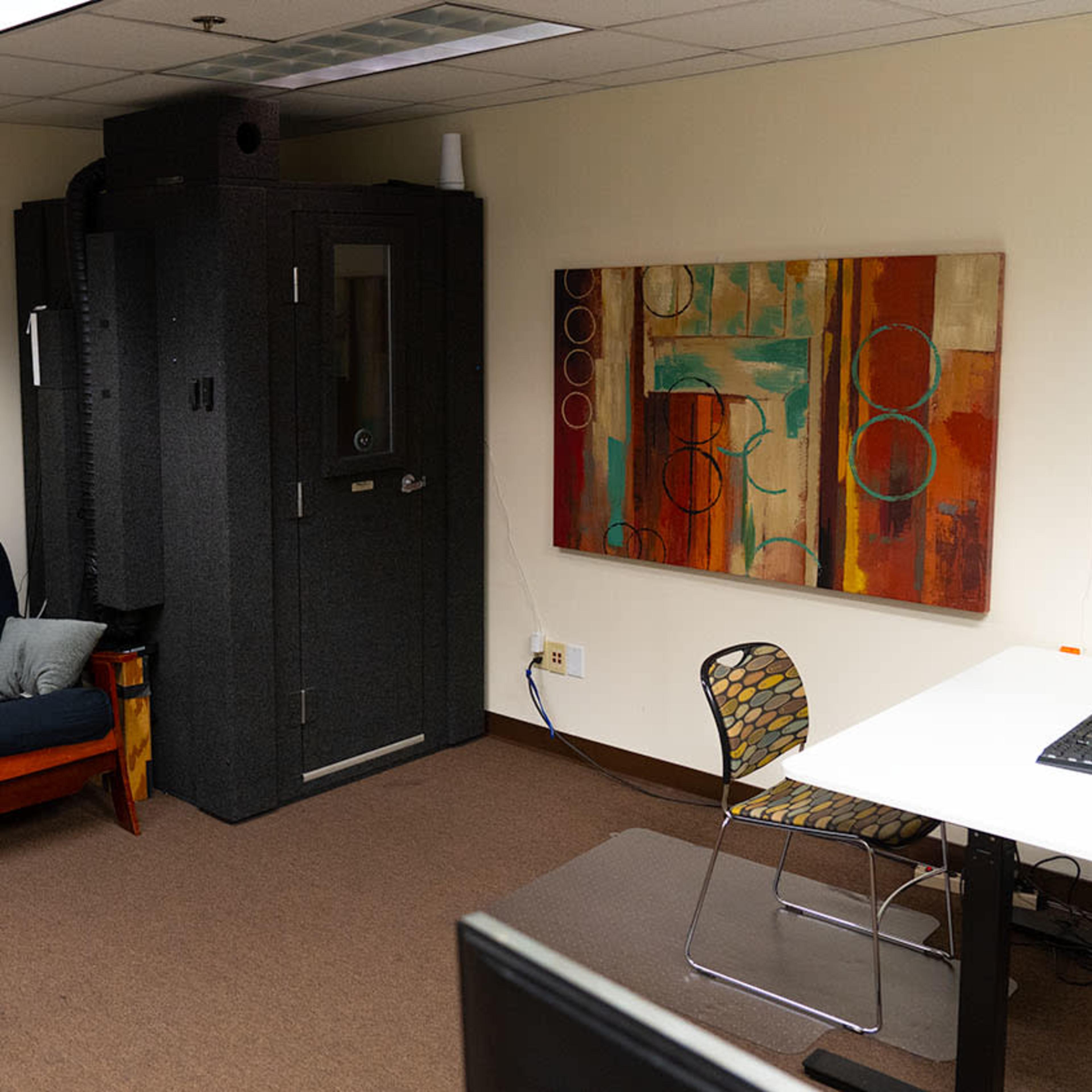 A small office space featuring a dark couch, a soundproof booth, an abstract painting on the wall, and a desk with a computer and a chair.