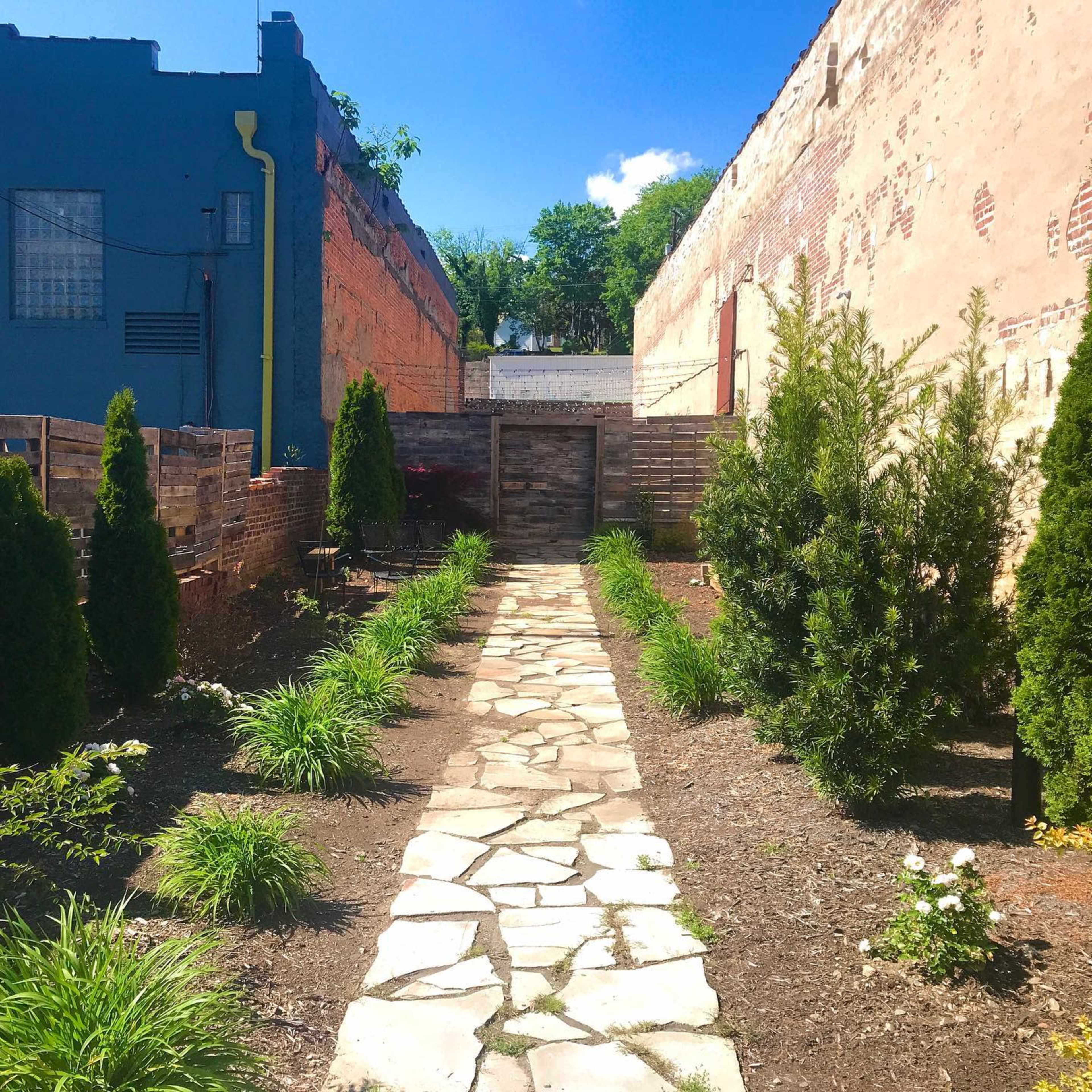 A stone pathway leads through a garden lined with small evergreen shrubs and plants, flanked by brick walls on either side.