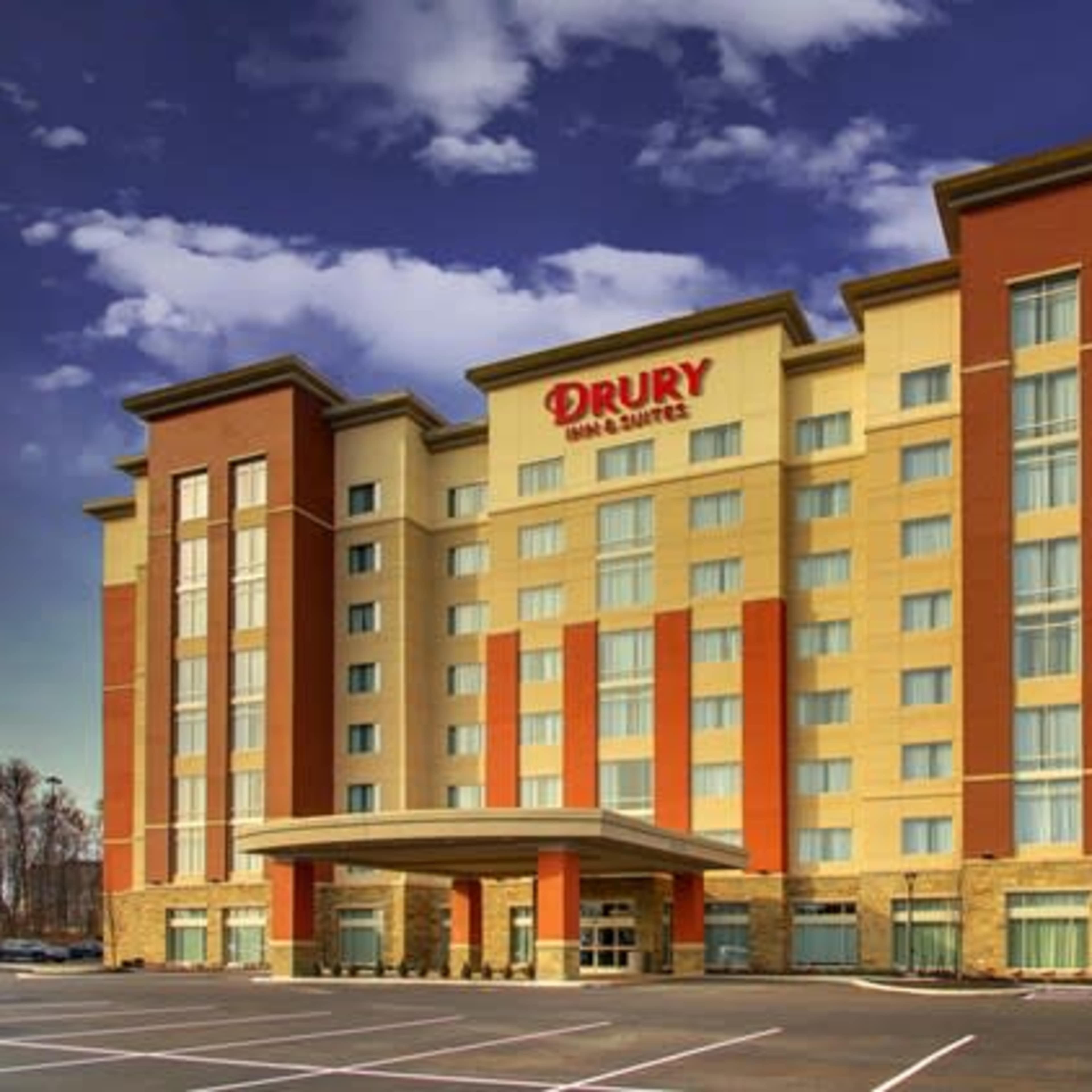 A six-story hotel building with a modern design and a large entrance, located in an open parking lot under a blue sky.