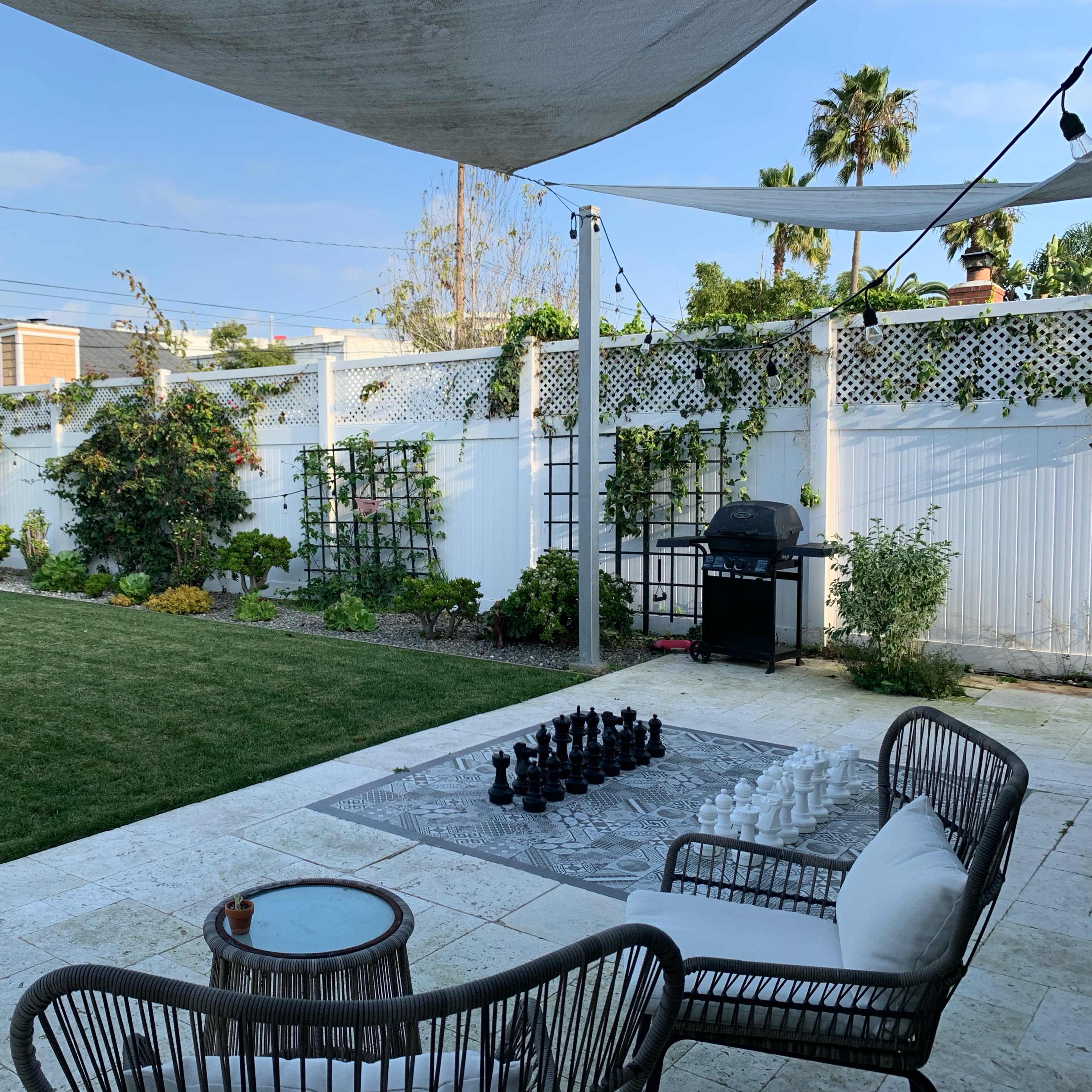 A spacious backyard features a large chess set on a patterned rug, surrounded by green grass, plants, and patio furniture under a shaded area.