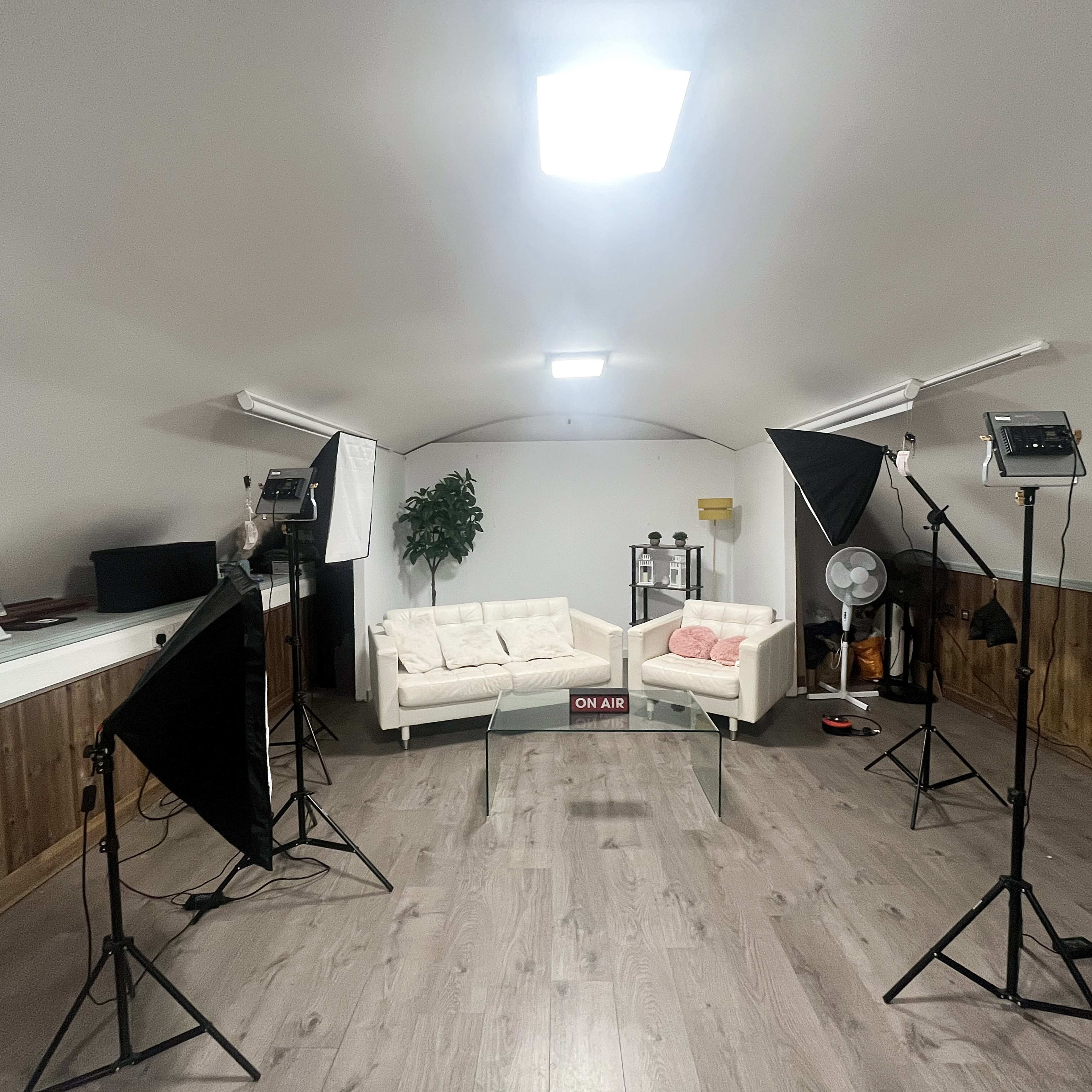 An indoor studio setup with two white sofas, lighting equipment, and a fan arranged in a spacious room.