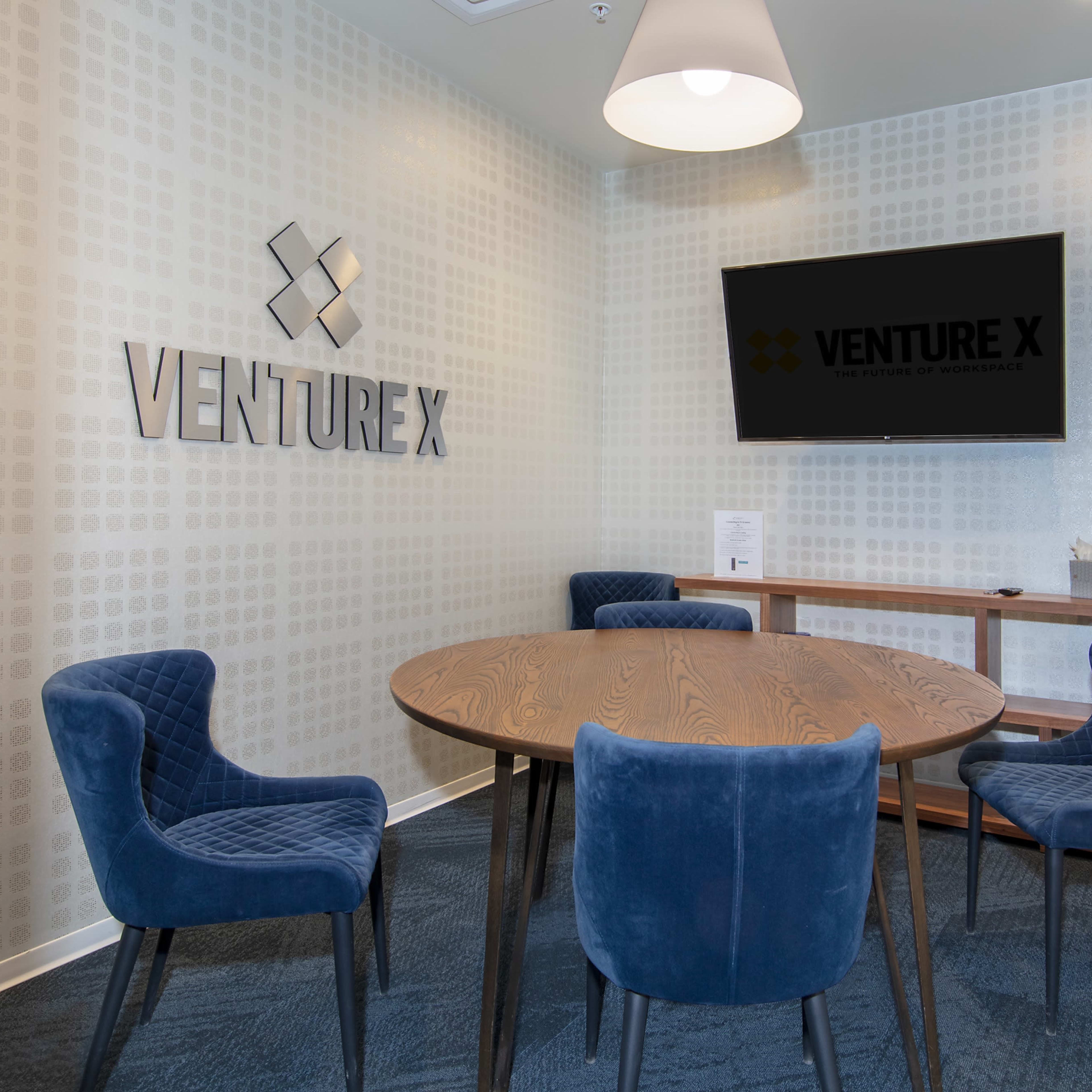 A modern conference room featuring a round wooden table surrounded by four blue upholstered chairs, with a large screen mounted on the wall displaying the "VENTURE X" logo.