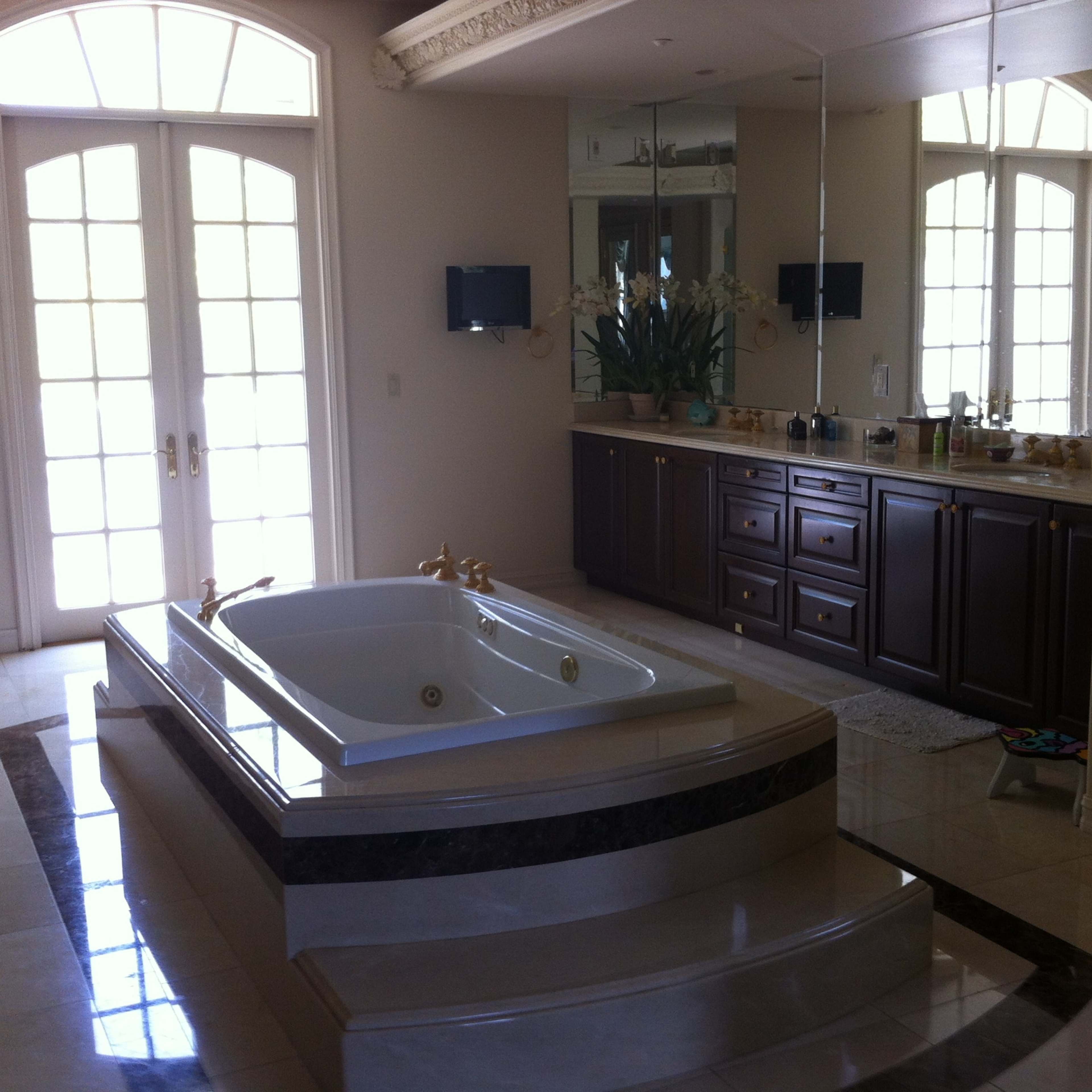 A spacious bathroom with a large bathtub at the center, two wall-mounted televisions, and a double vanity against a mirrored wall.