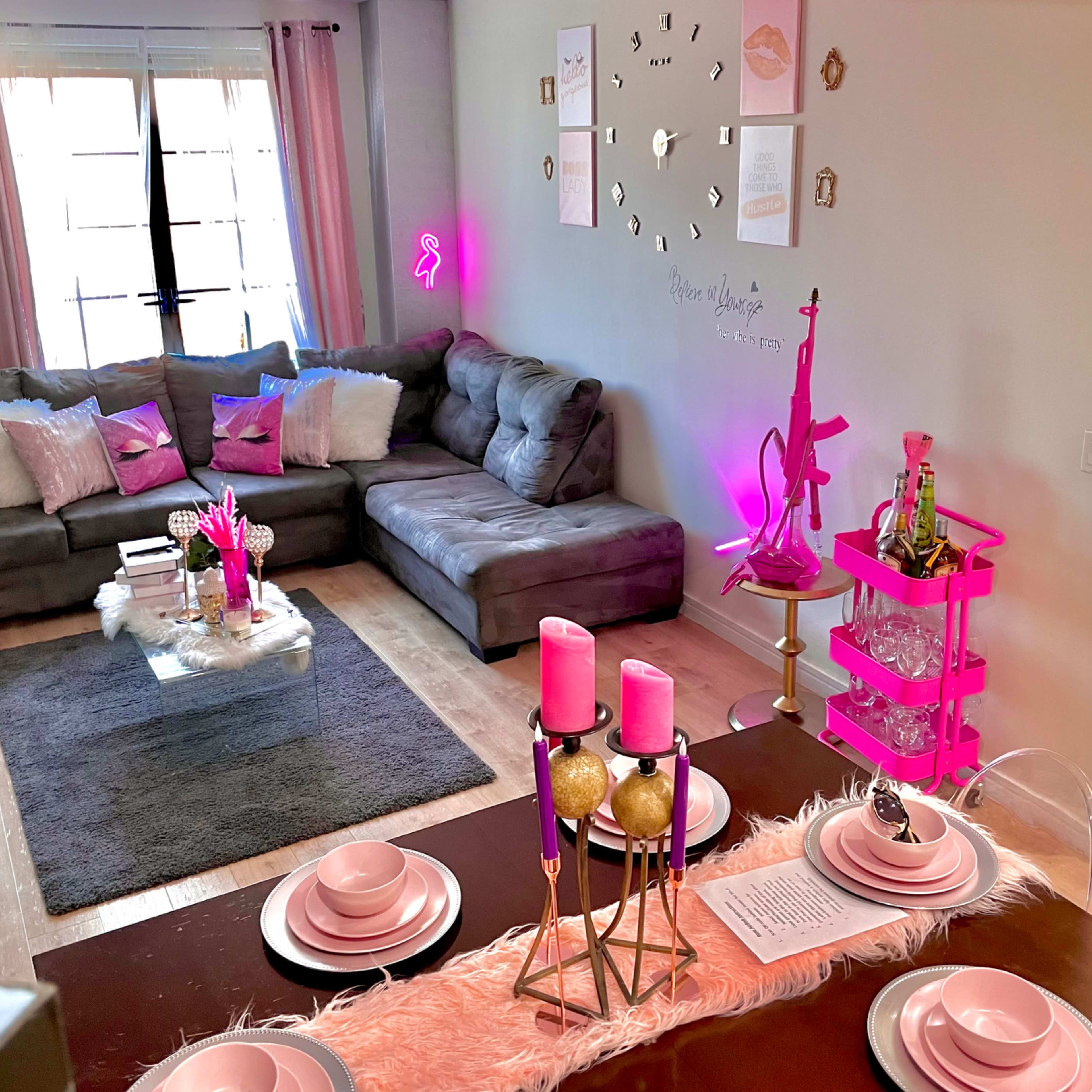 A modern living space with a gray sectional sofa, a dining table set with pink decor and candles, and decorative elements like a pink cart and wall art.