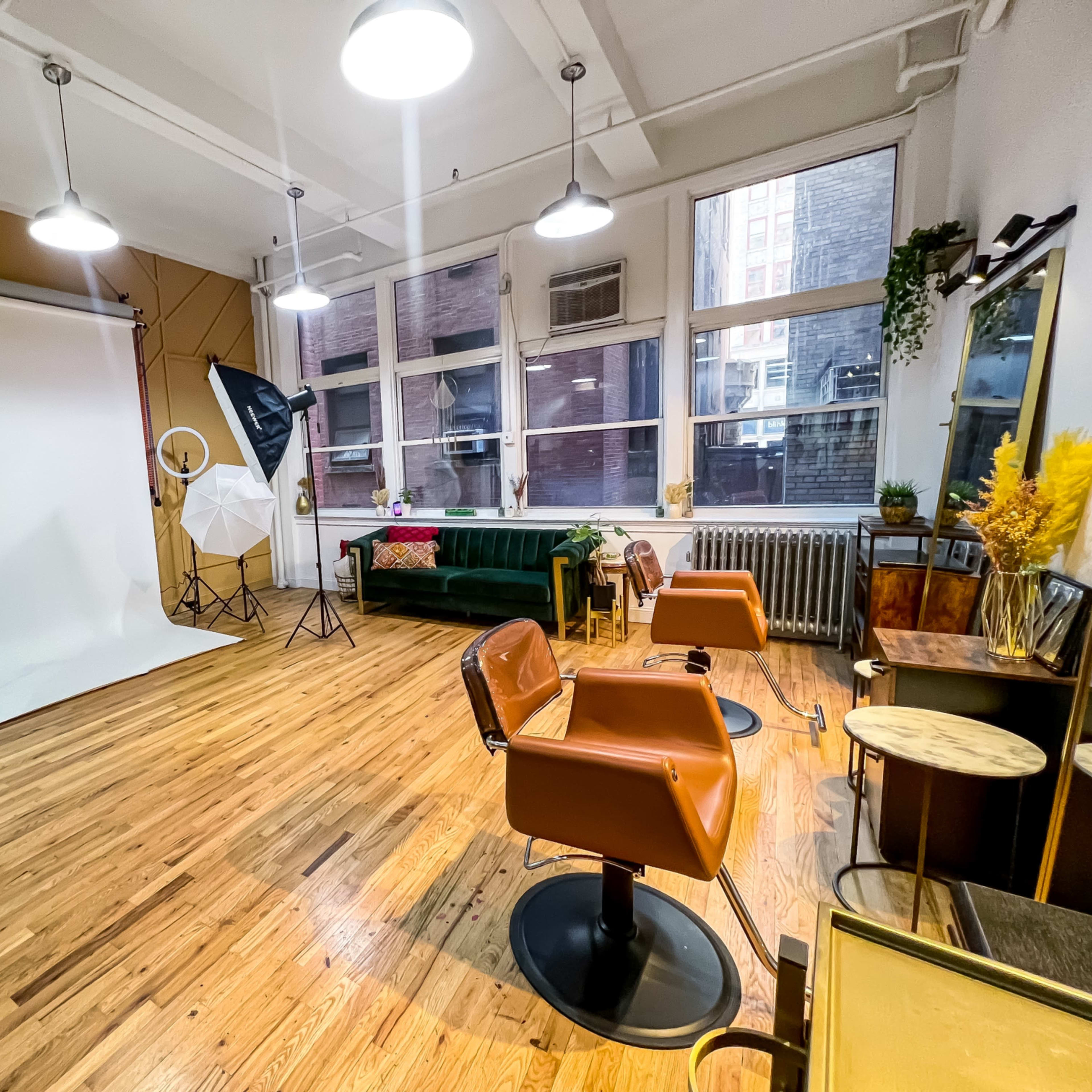 A spacious studio setup with a white backdrop, two softbox lights, a green couch, and several salon chairs on a wooden floor.