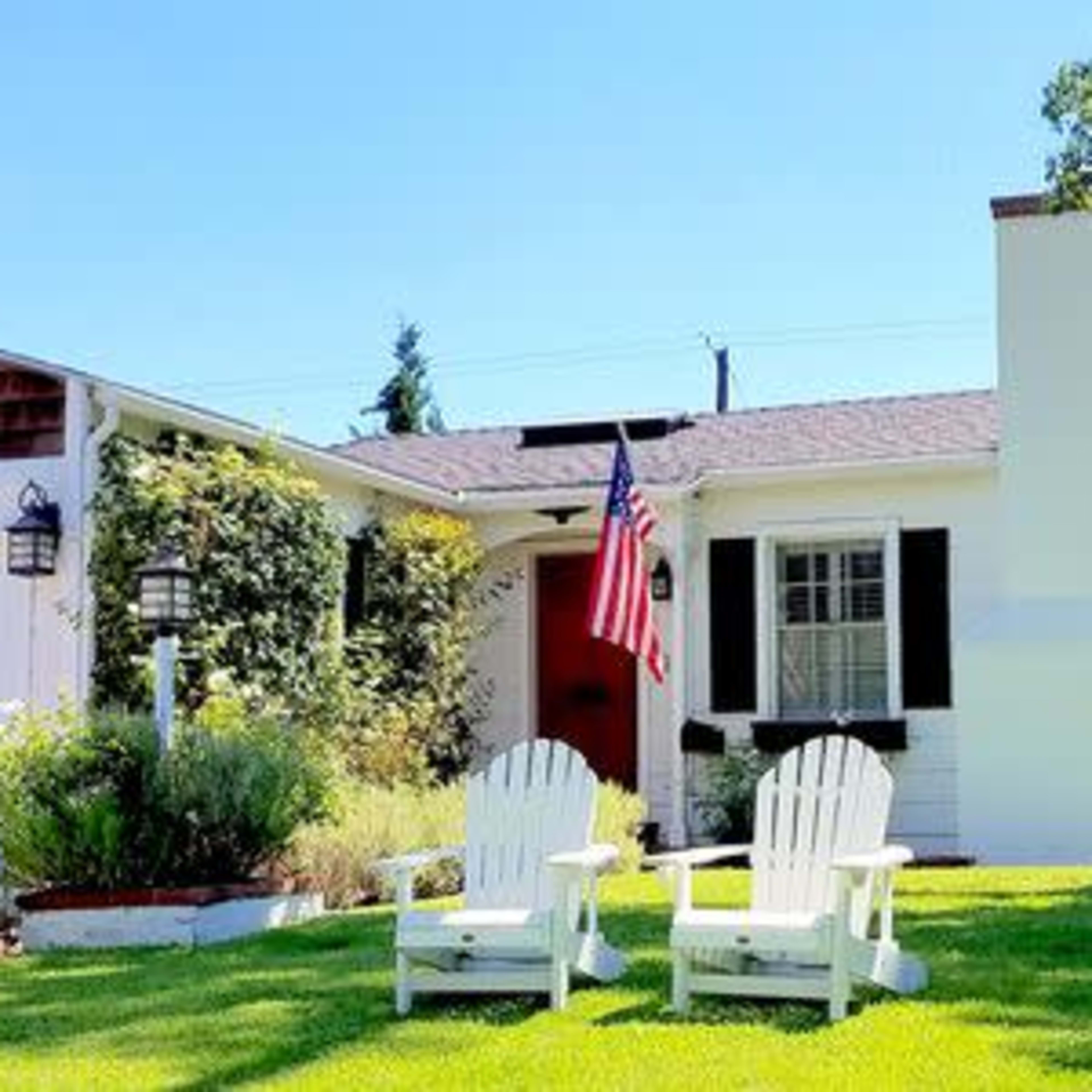 A white house with black shutters and an American flag stands in front of a well-manicured lawn featuring two white adirondack chairs.