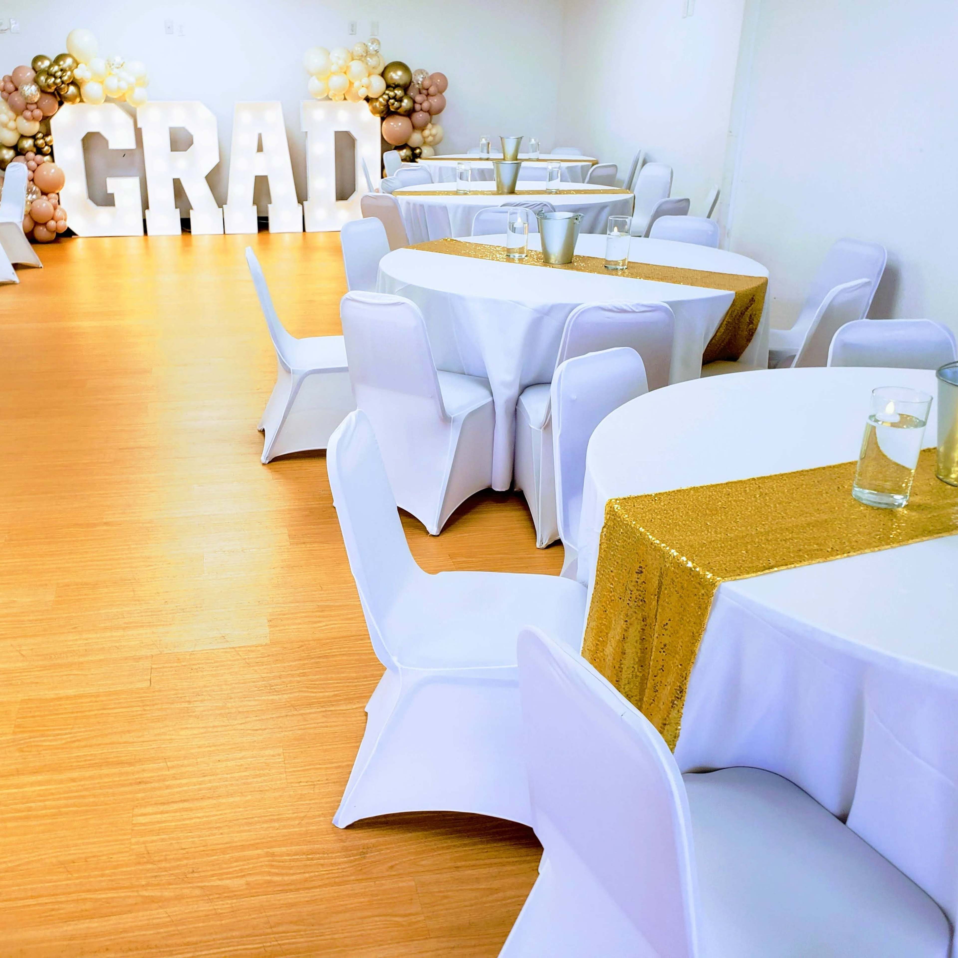 A decorated event space with white tables, white chairs, and a "GRAD" display made of large letters surrounded by balloon decorations.