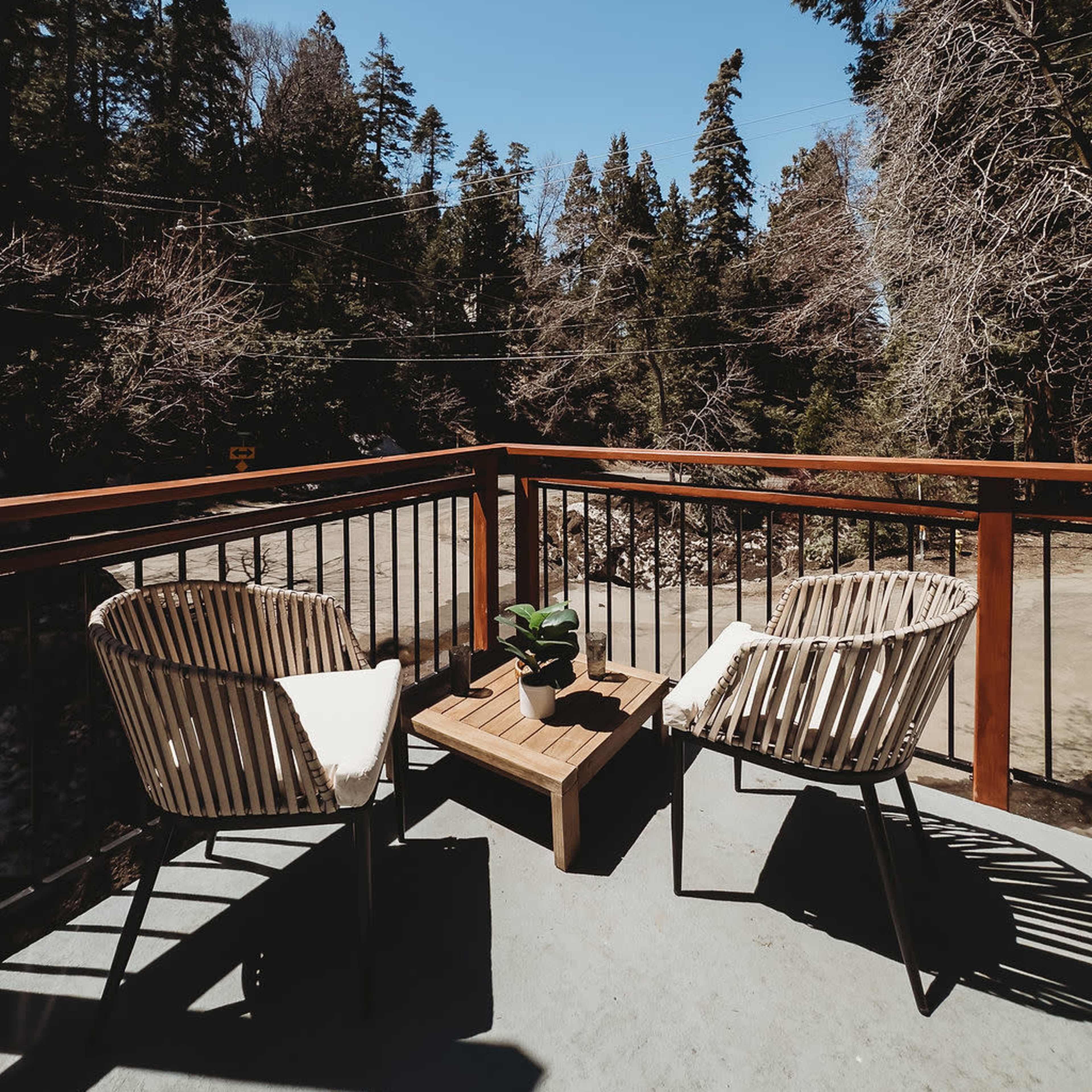 A wooden deck features two chairs and a small table, overlooking a wooded area under a clear blue sky.