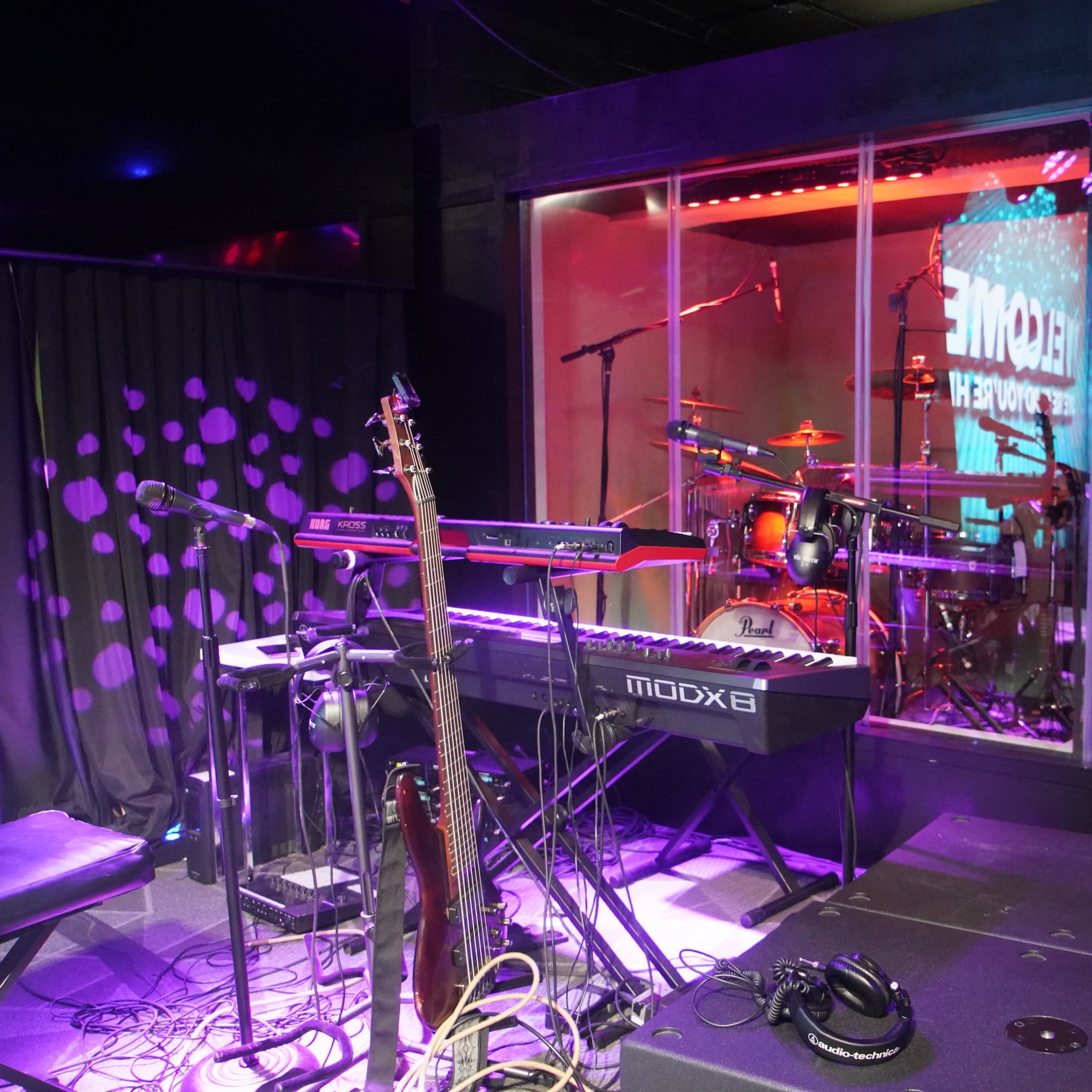 A music performance setup features a keyboard, microphone, and drum kit inside a glass enclosure, with stage lighting creating an atmospheric scene.