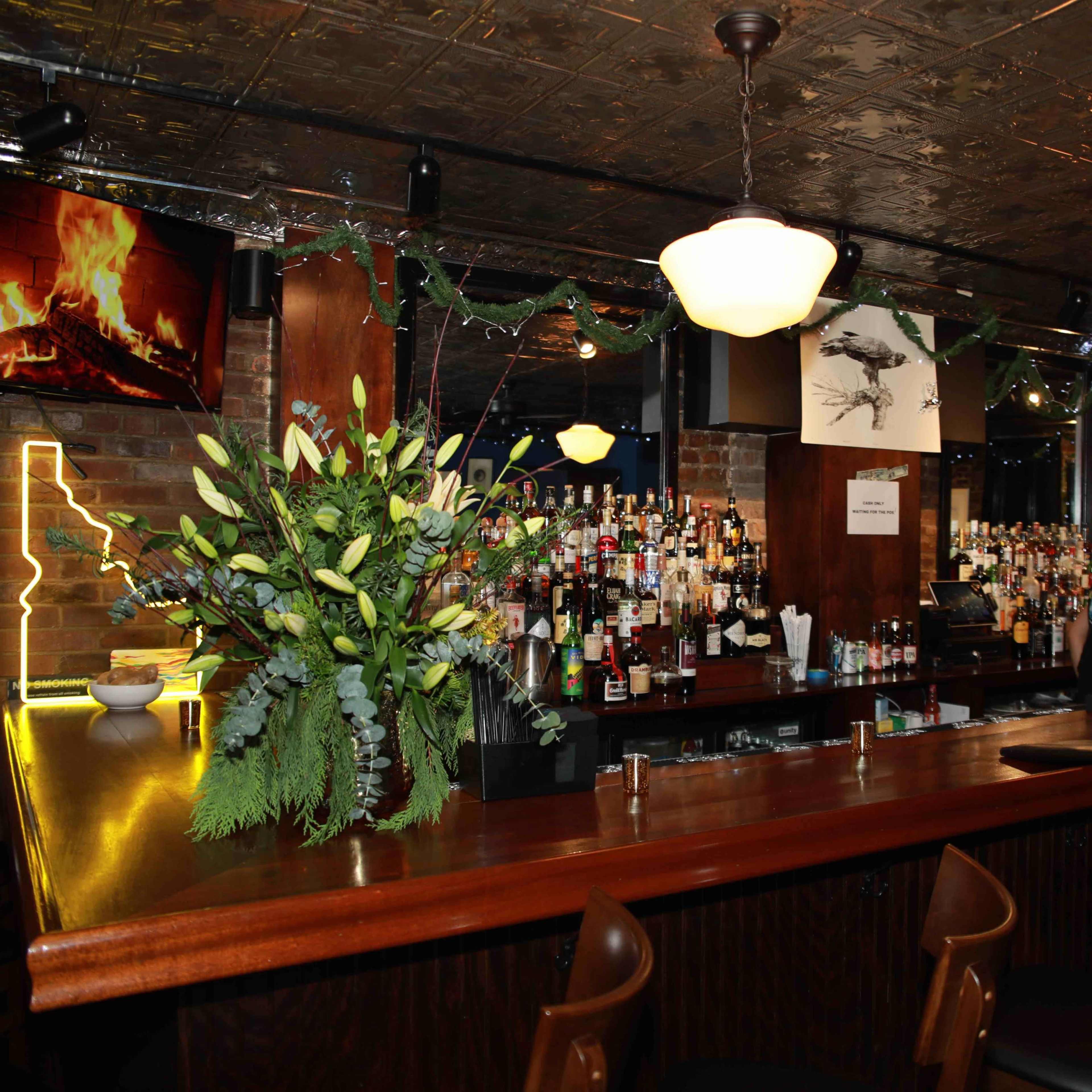 A bar with a wooden counter, a variety of alcoholic beverages on display, and decorations including a fireplace on a screen and a floral arrangement.