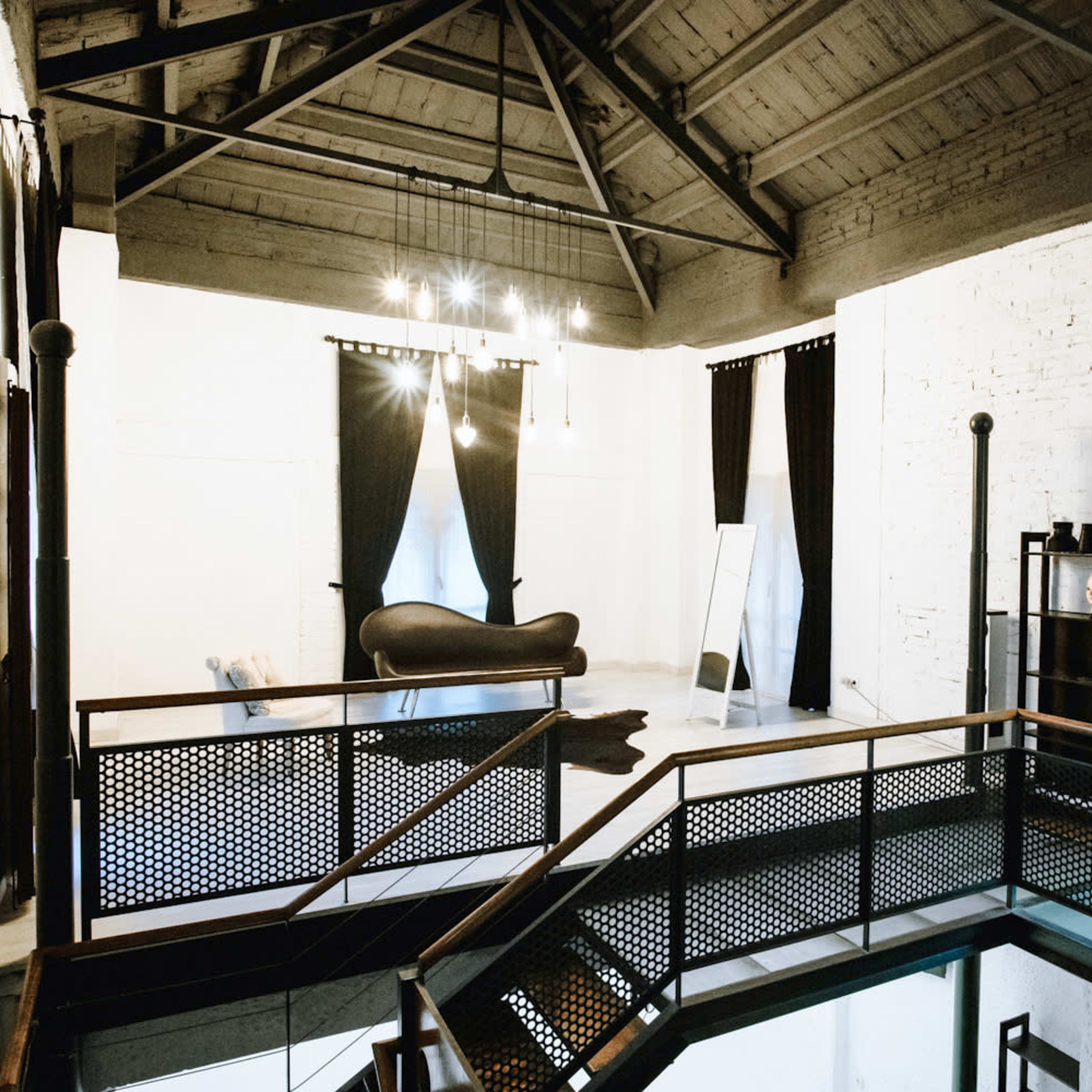A spacious interior with a high ceiling, featuring a balcony overlooking a room with a dark chaise lounge and large windows covered by black curtains.