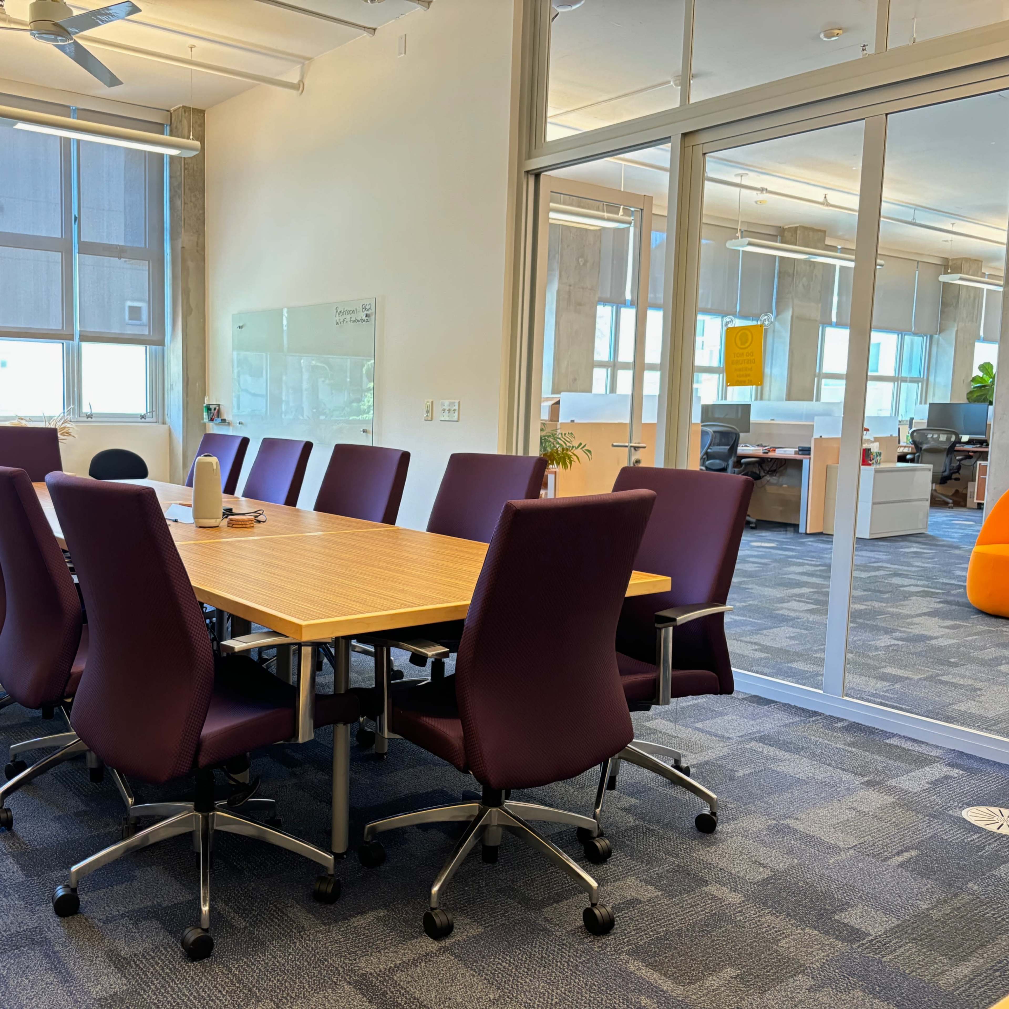 A conference room features a large wooden table surrounded by twelve purple chairs, with large windows allowing natural light.
