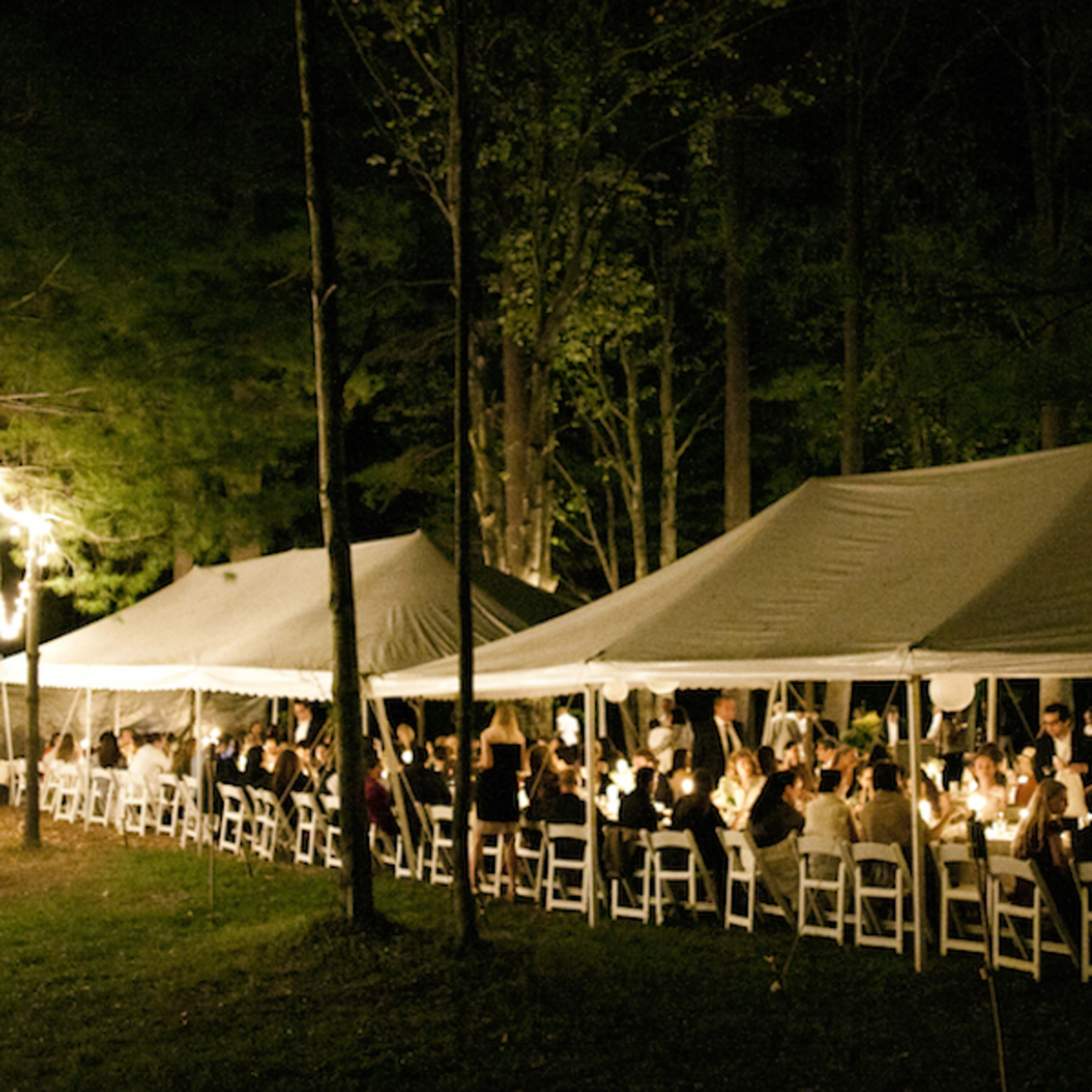 A group of people gather under string lights at tables arranged beneath large tents in a forested setting at night.