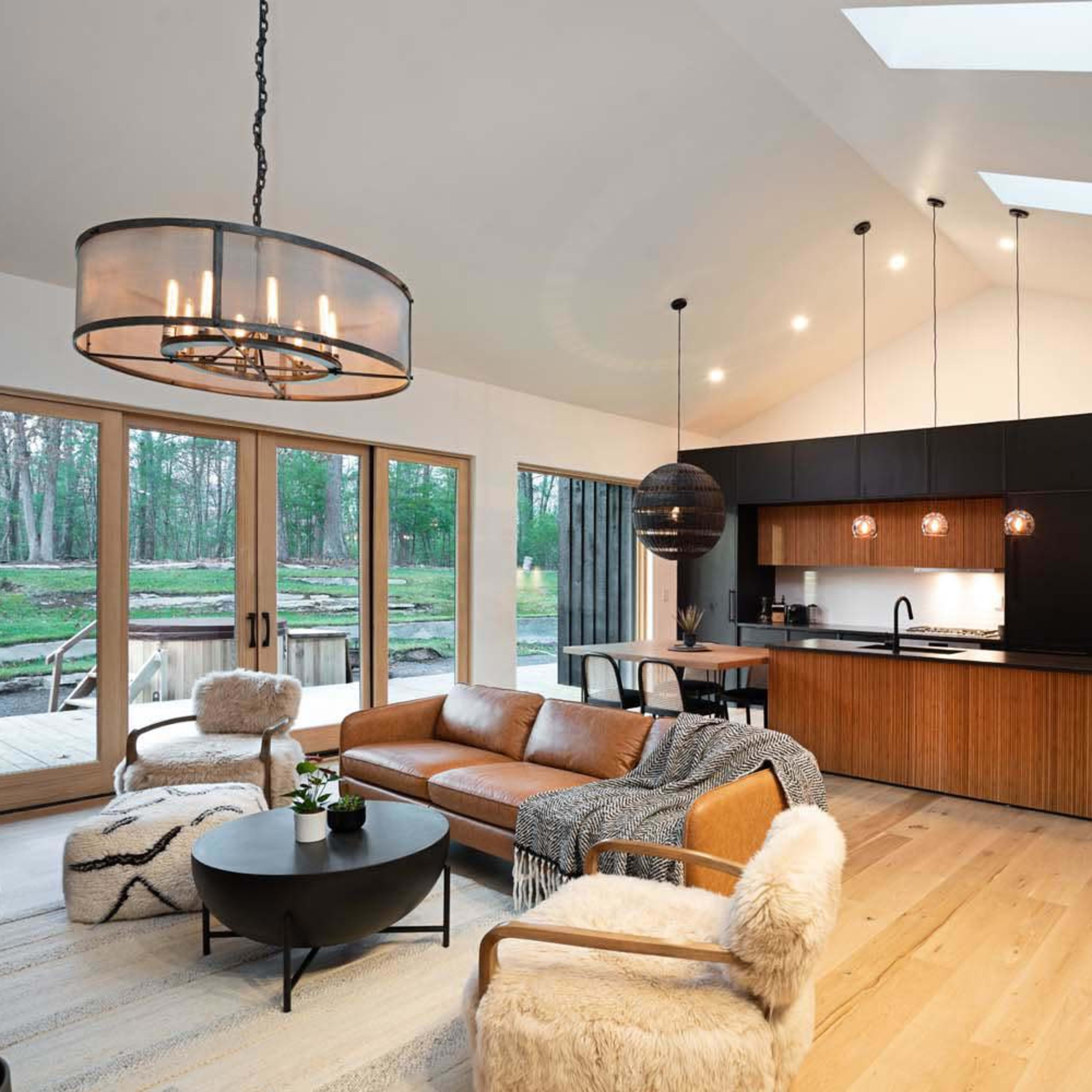 A modern living room with a leather sofa, a round coffee table, and a kitchen area featuring dark cabinetry and large windows that open to a wooded outdoor space.