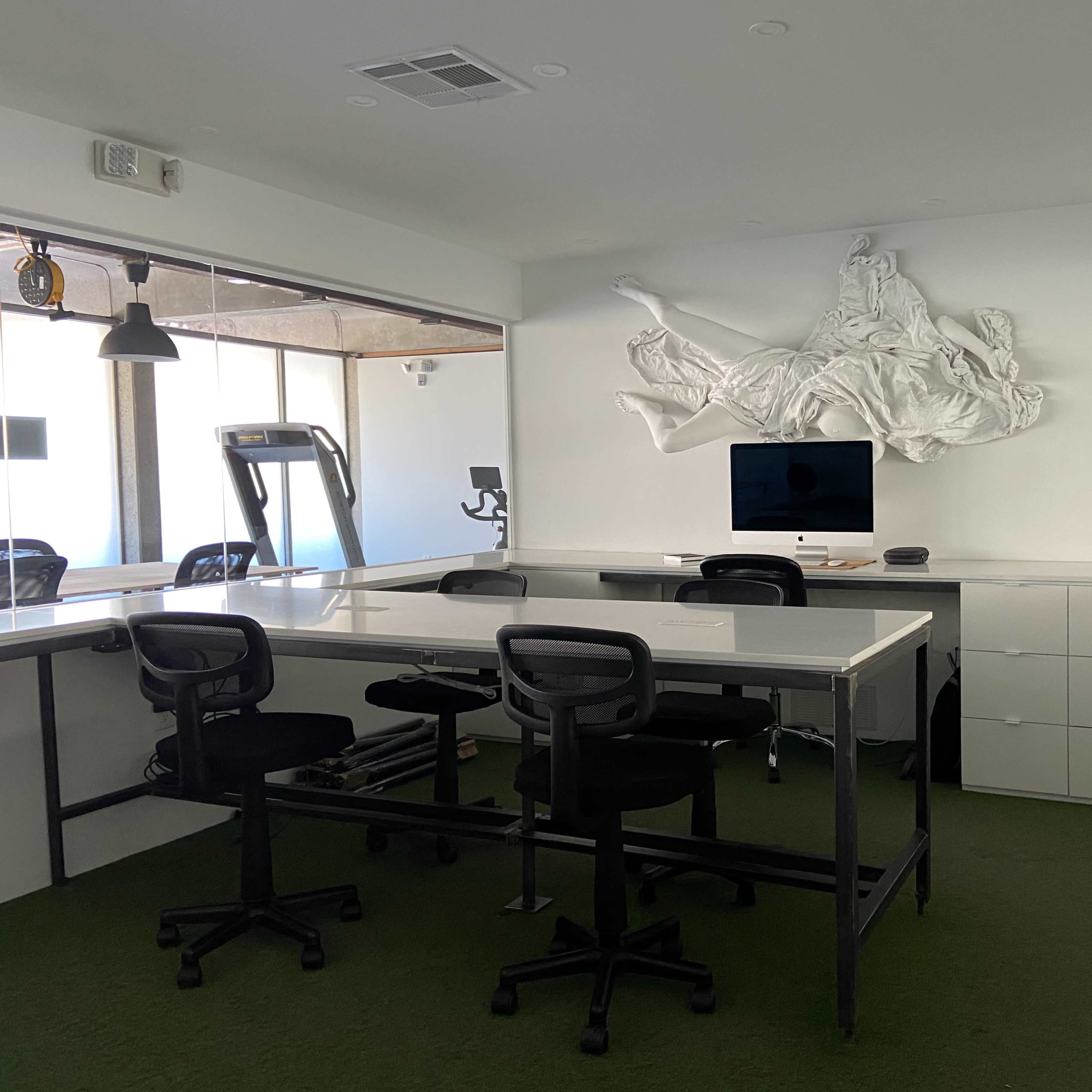 A modern office space features a large white wall sculpture, a desk with chairs, and a treadmill in the background.