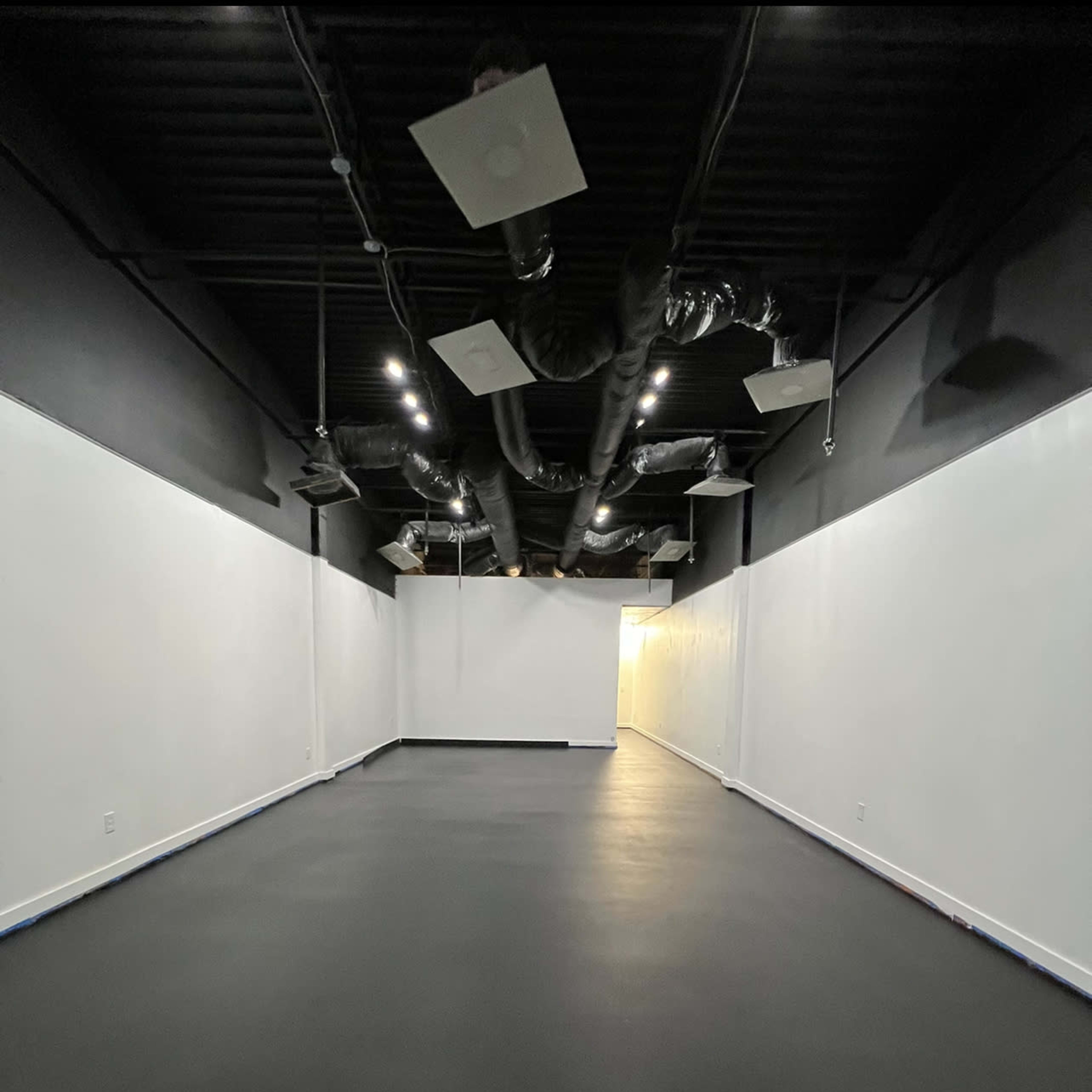 An empty room with black flooring and white walls, featuring exposed ductwork and overhead lighting.
