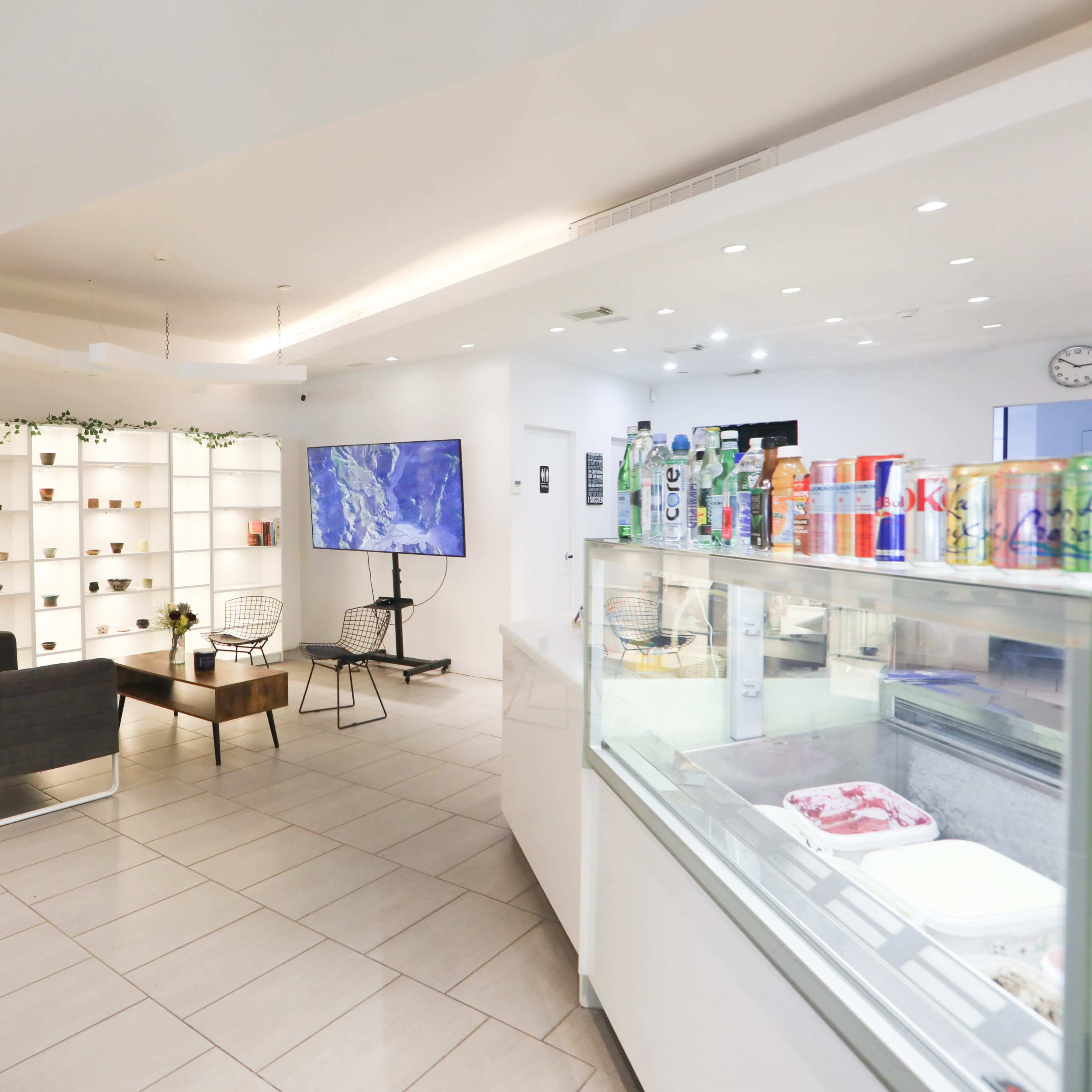 A modern café interior with a seating area, a display case for desserts, a large screen, and shelves showcasing various items.