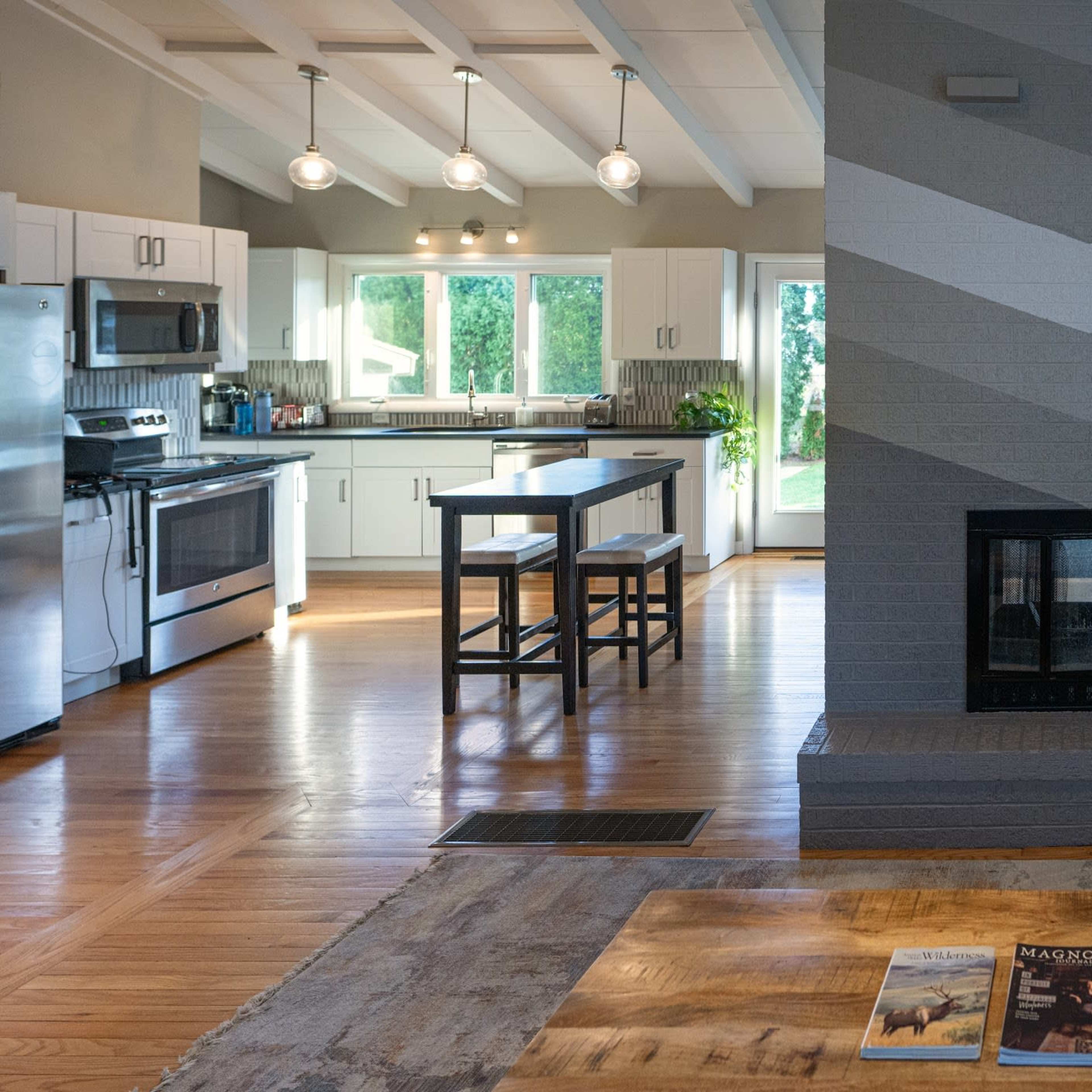 A modern kitchen with stainless steel appliances and a geometric-patterned fireplace, featuring a wooden dining table and magazines on the coffee table.