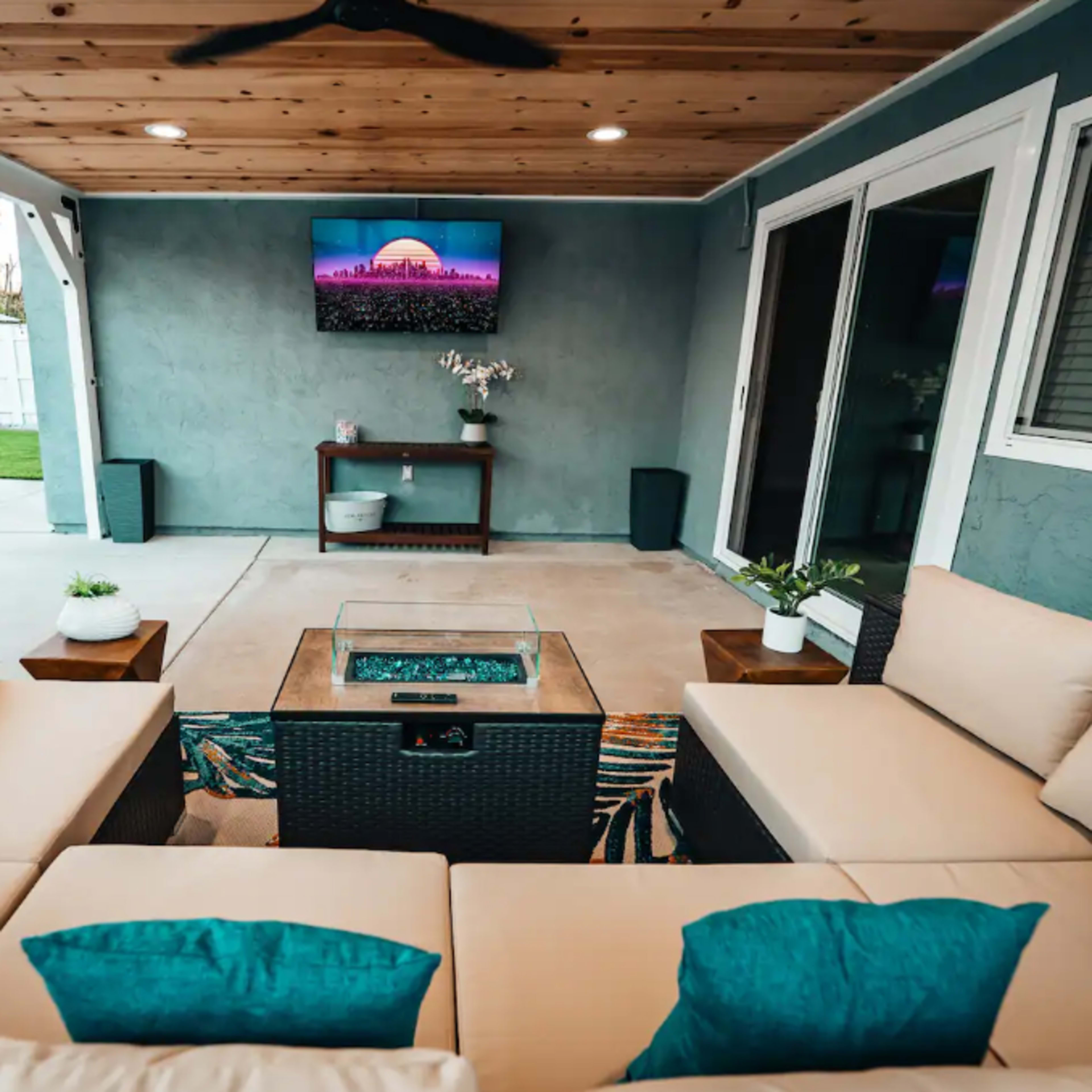 A covered outdoor living space features a sectional sofa with blue cushions, a fire pit table, and a wall-mounted television.