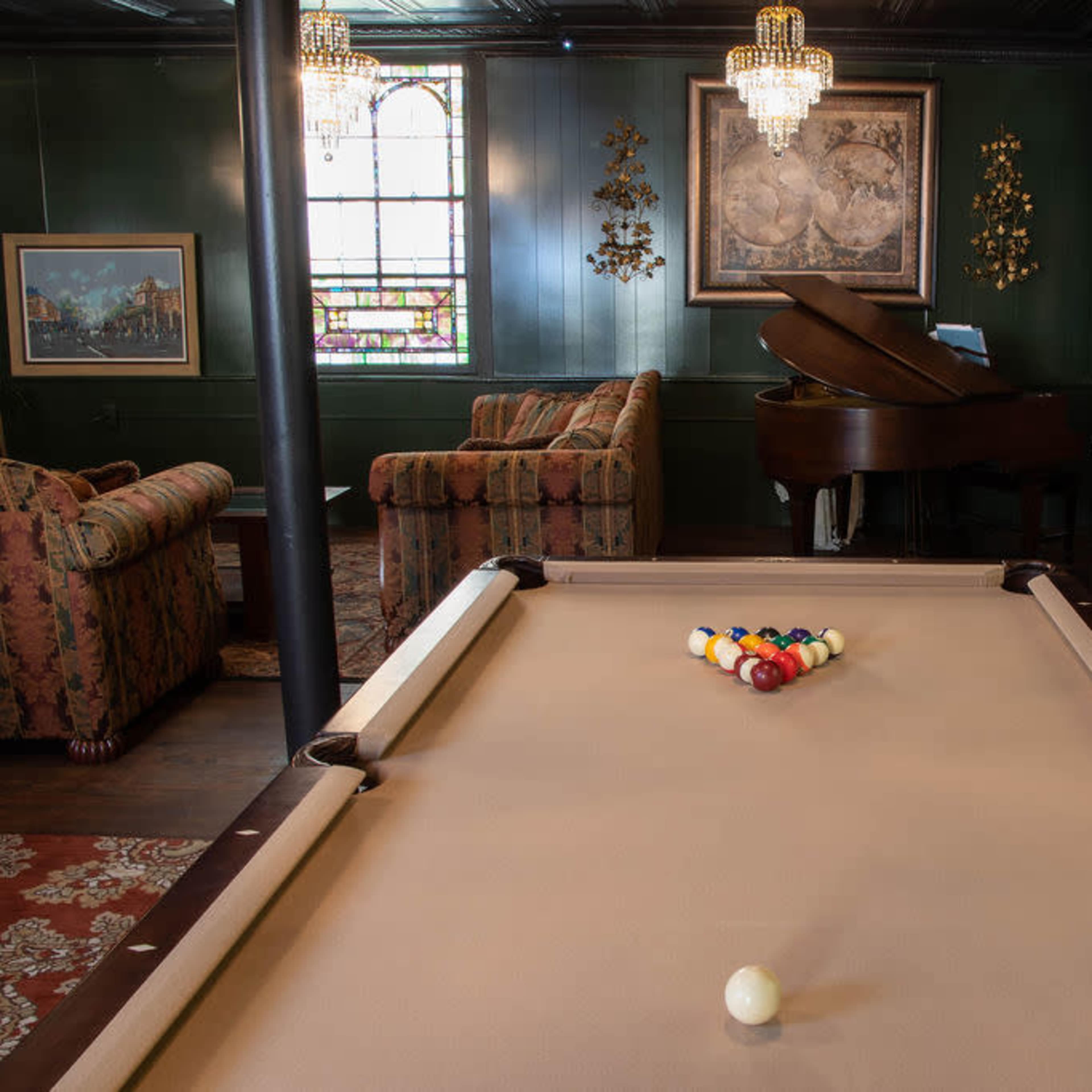 A room with a billiard table in the foreground, a grand piano nearby, and cozy seating arrangements against green walls, illuminated by chandeliers.