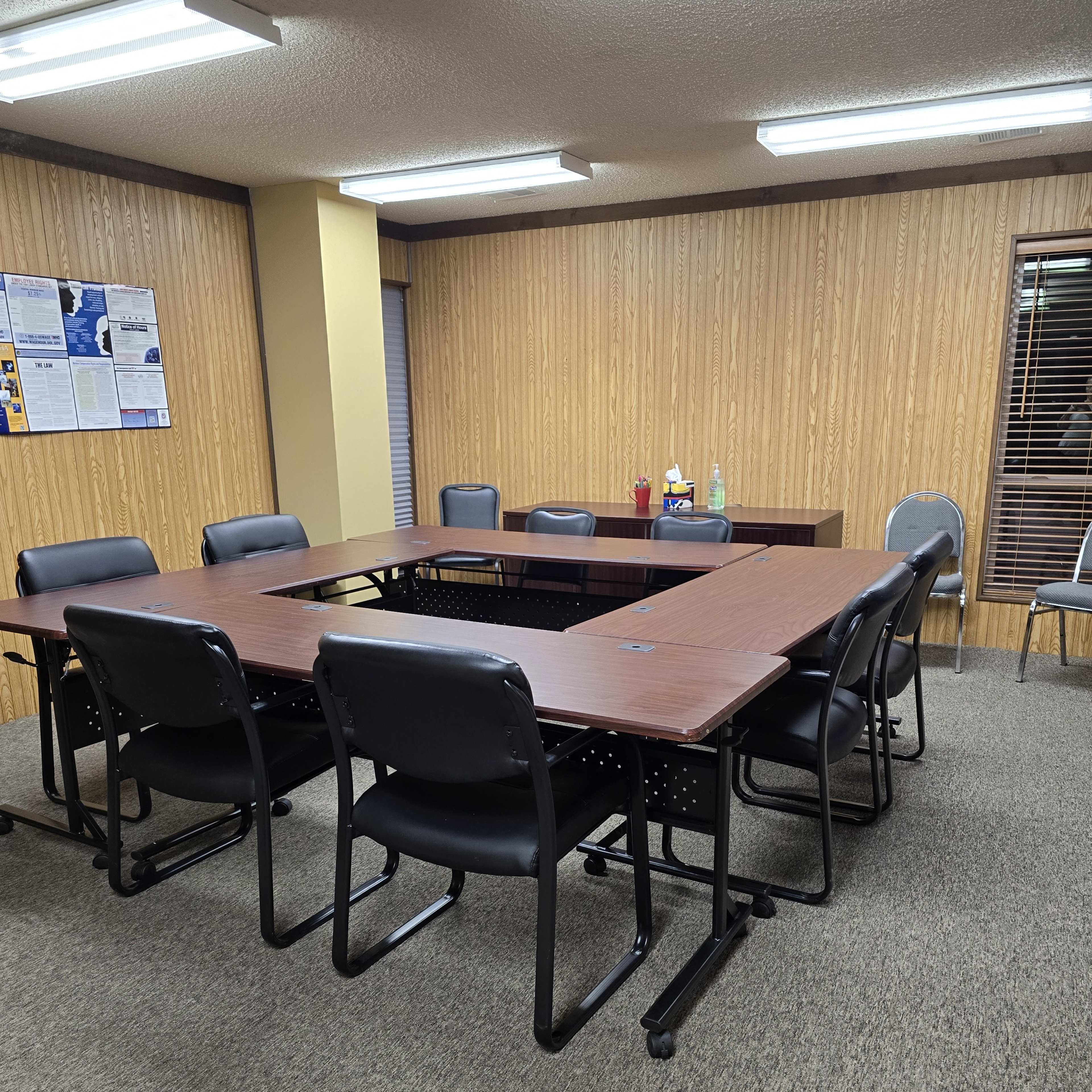 A rectangular conference room table surrounded by black chairs is positioned in a room with wood-paneled walls and large windows.