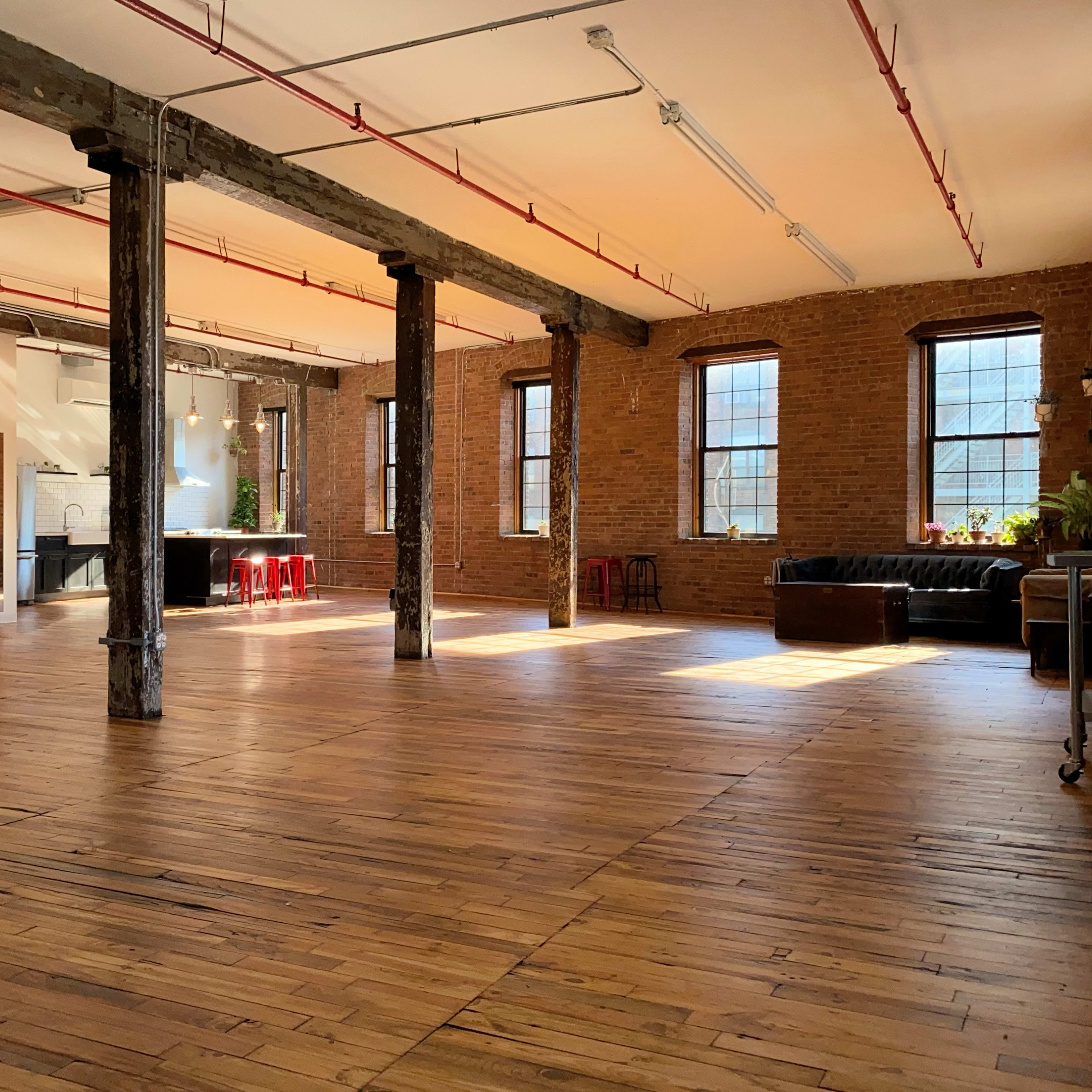 An open industrial space with exposed brick walls, wooden floors, large windows, and various seating arrangements.