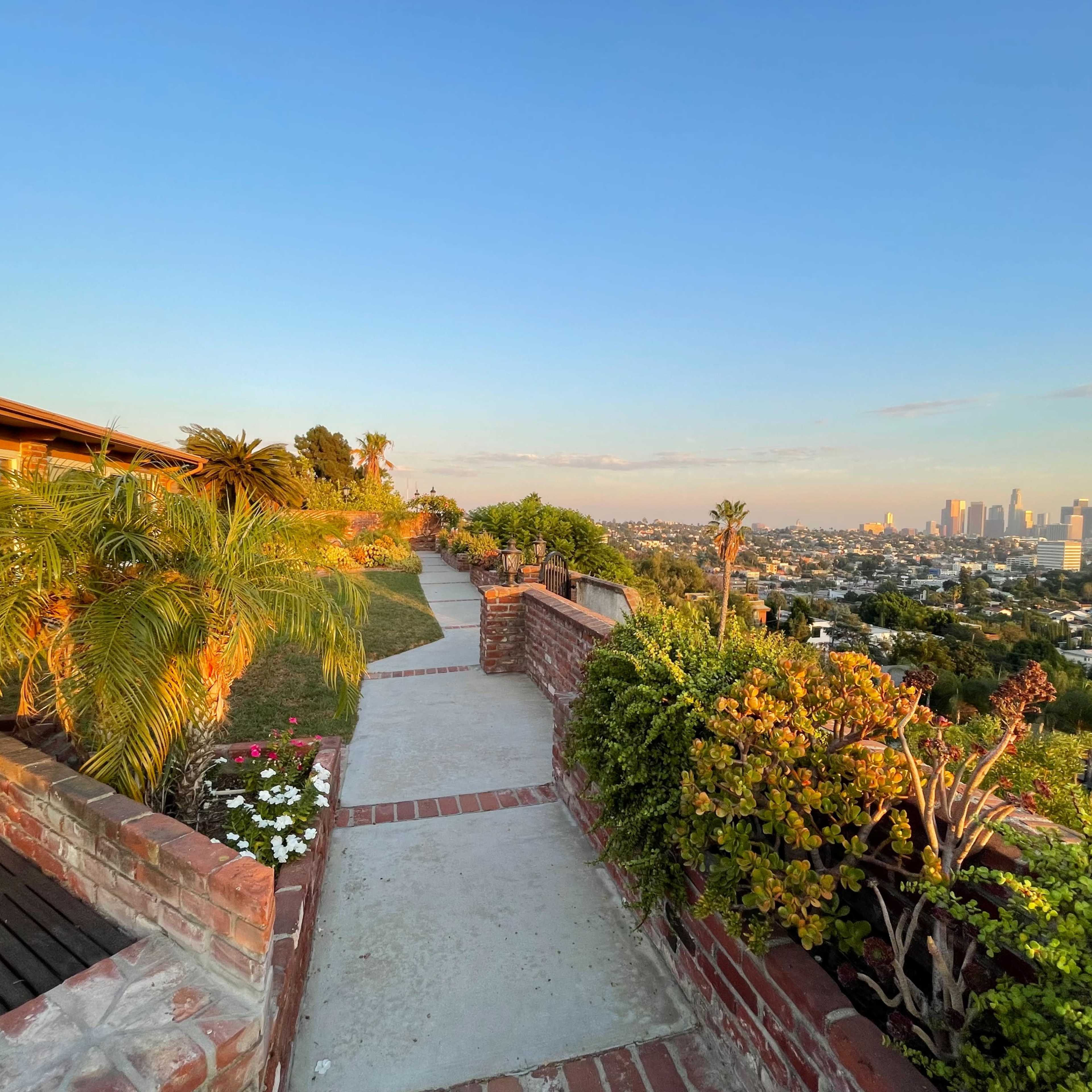 A pathway lined with plants and flowers leads to a view of the Los Angeles skyline from a hilltop residence.