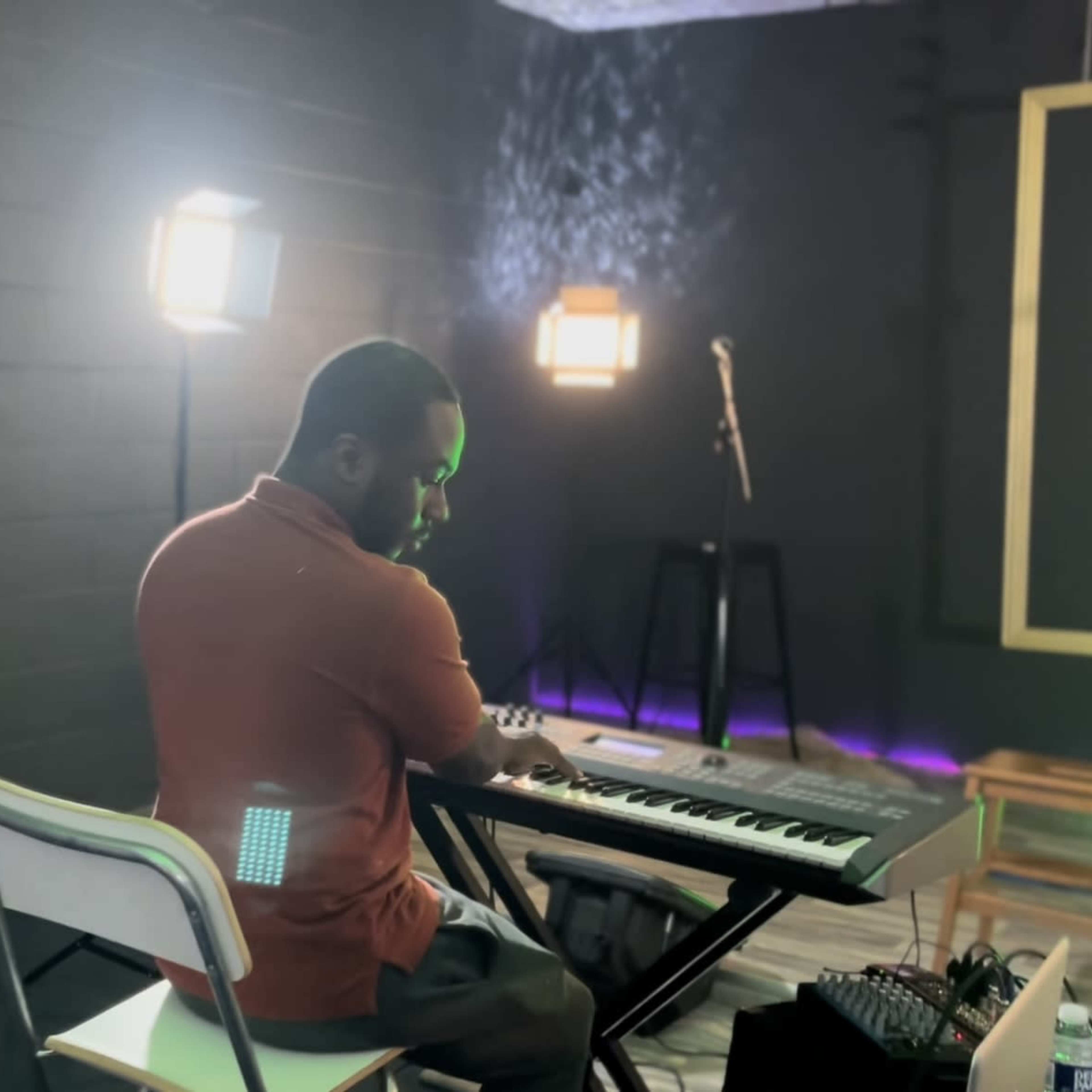 A musician plays a keyboard in a dimly lit room with soft lighting and empty chairs nearby.