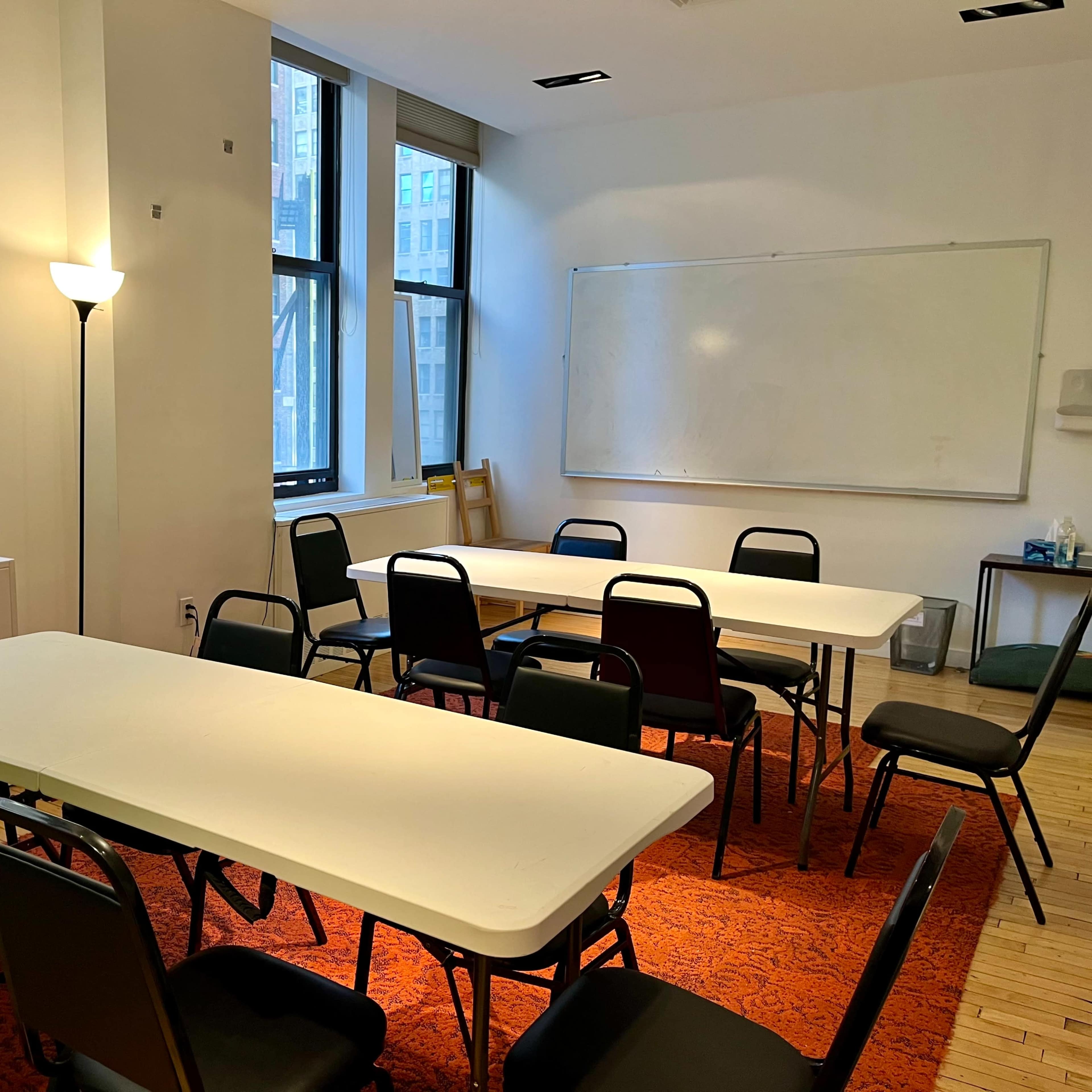 A large meeting room features several rectangular tables surrounded by chairs, with large windows allowing natural light and a whiteboard mounted on the wall.