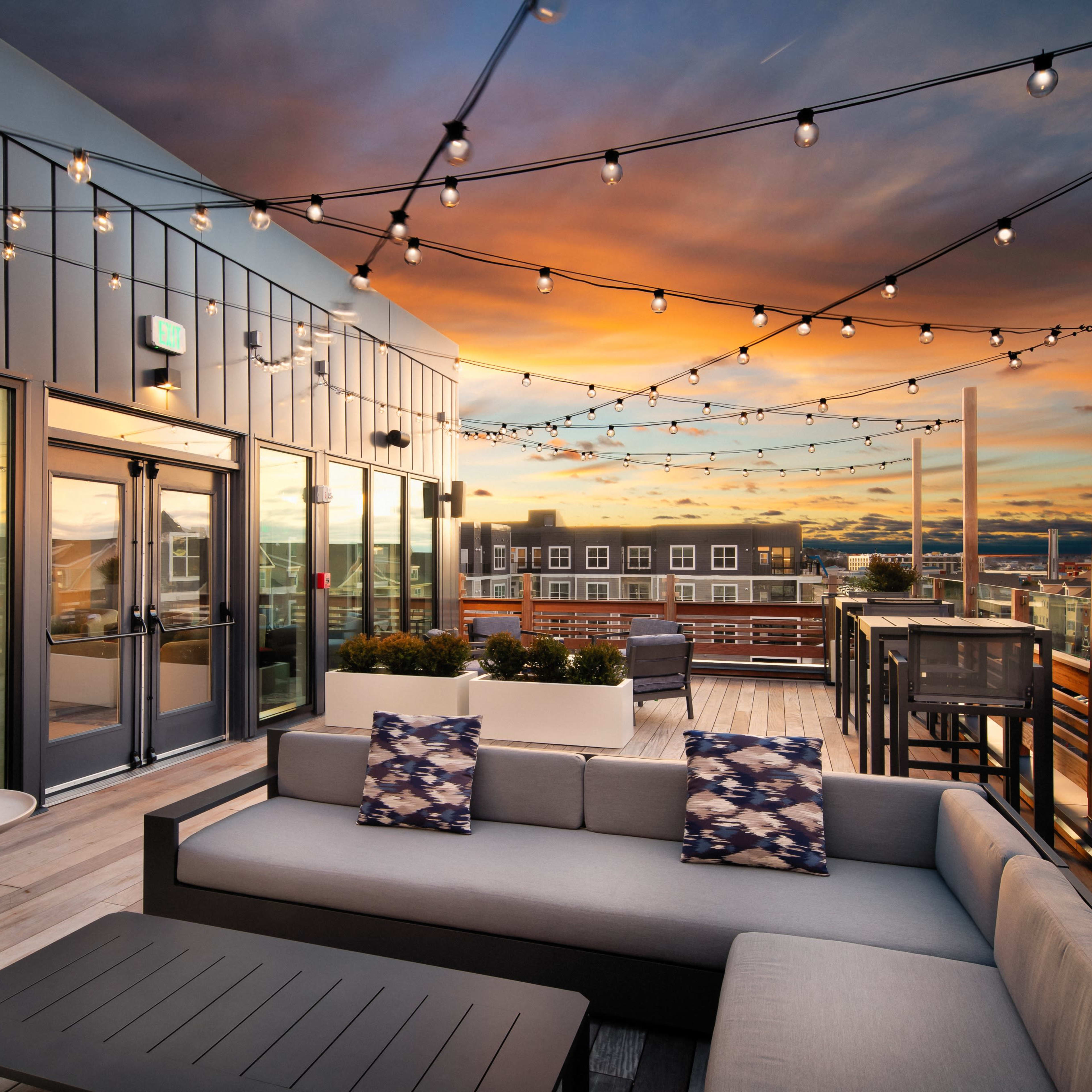 A rooftop terrace features a spacious sectional sofa, decorative cushions, and string lights overhead, with a view of a sunset sky and cityscape.