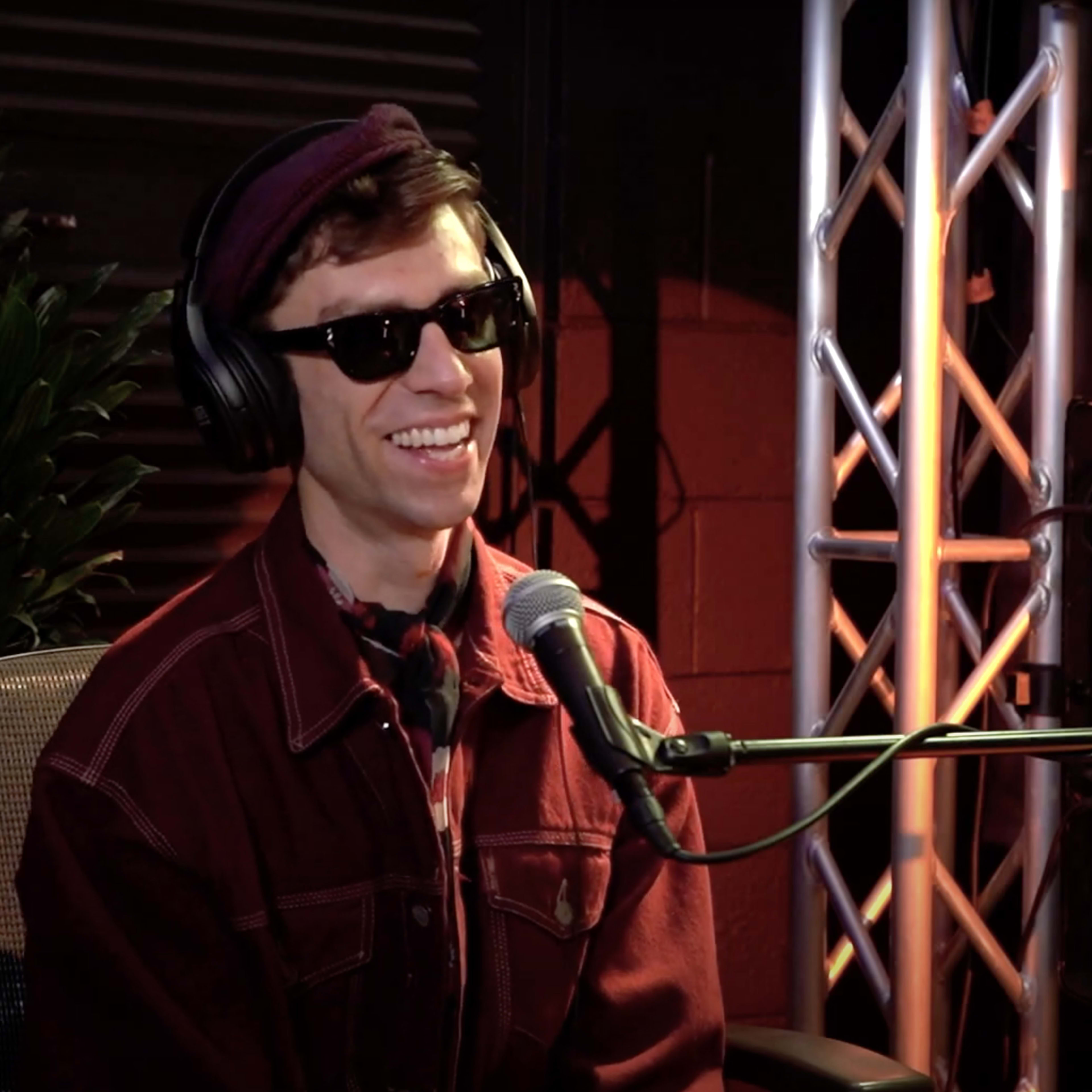 A man wearing sunglasses and headphones sits in front of a microphone, surrounded by lighting rigs and a plant.